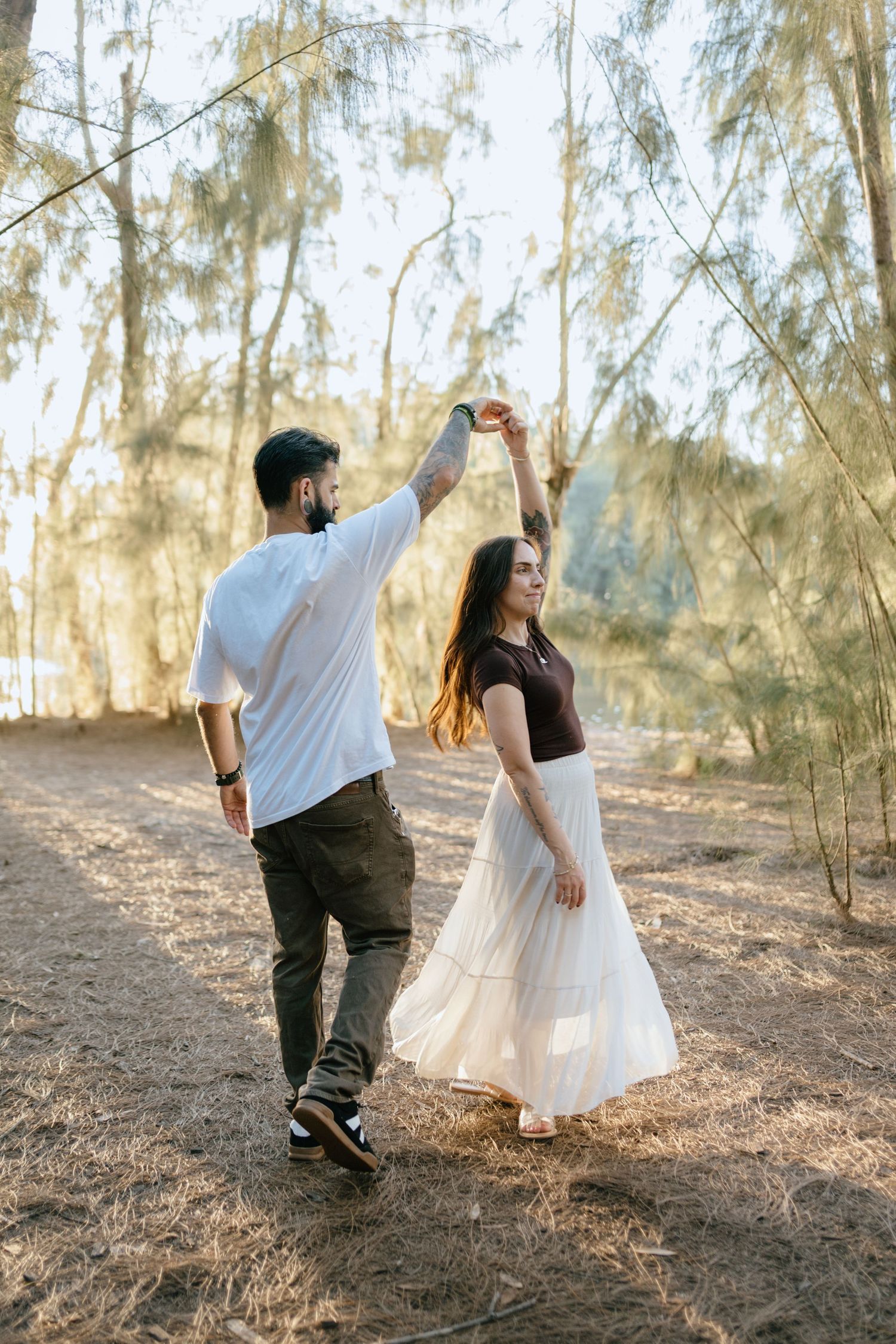 Man with tattoos twirls woman in white skirt among sunlit trees in a joyful outdoor dance.