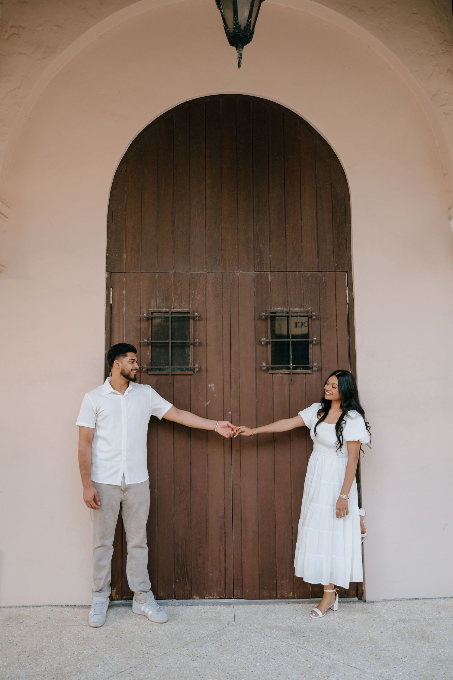 Couple in white outfits holding hands in front of a large arched wooden door.