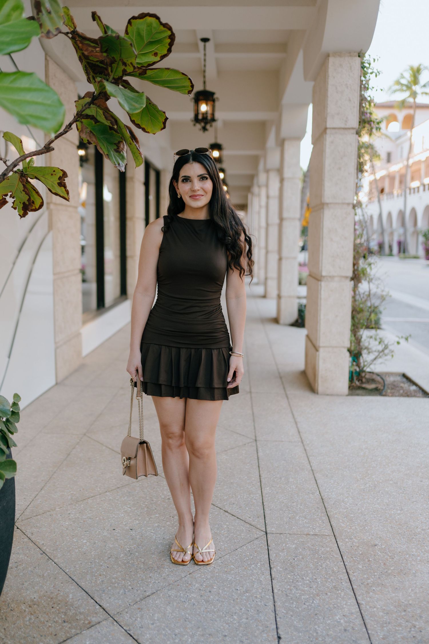 Young woman in black sleeveless mini dress with ruffled hem, holding beige bag, standing under colonnade.