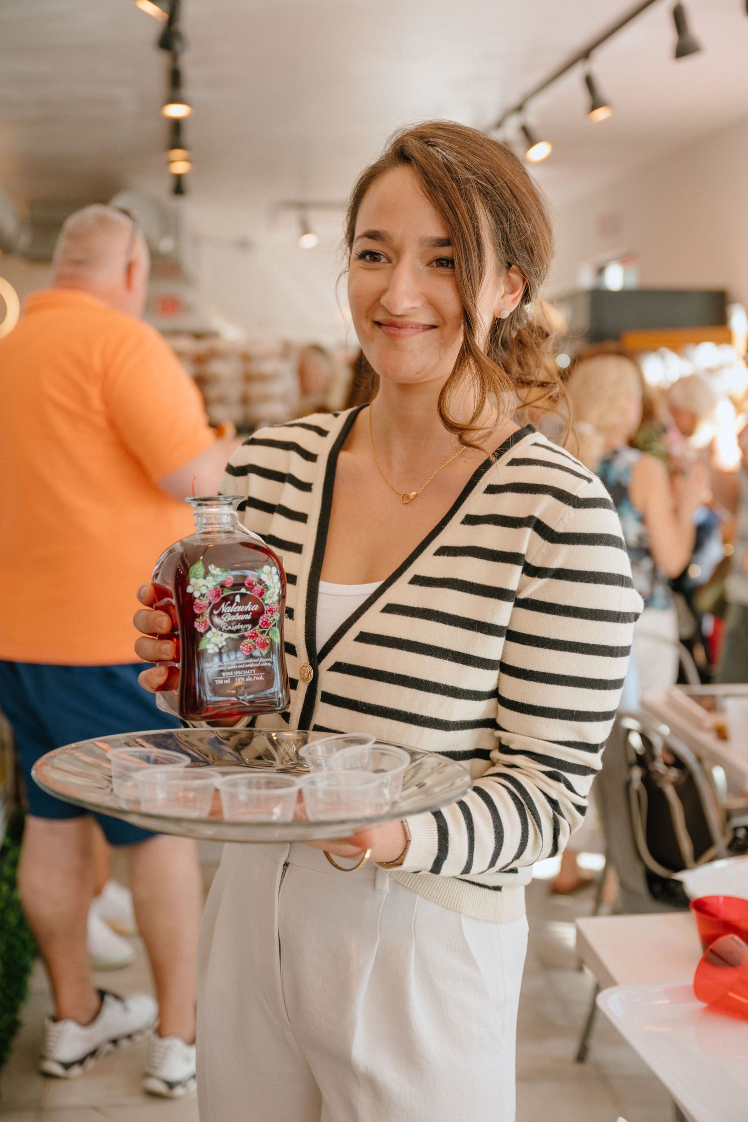 Smiling woman in striped shirt holding a decorative bottle and tray of sample cups at a food event.