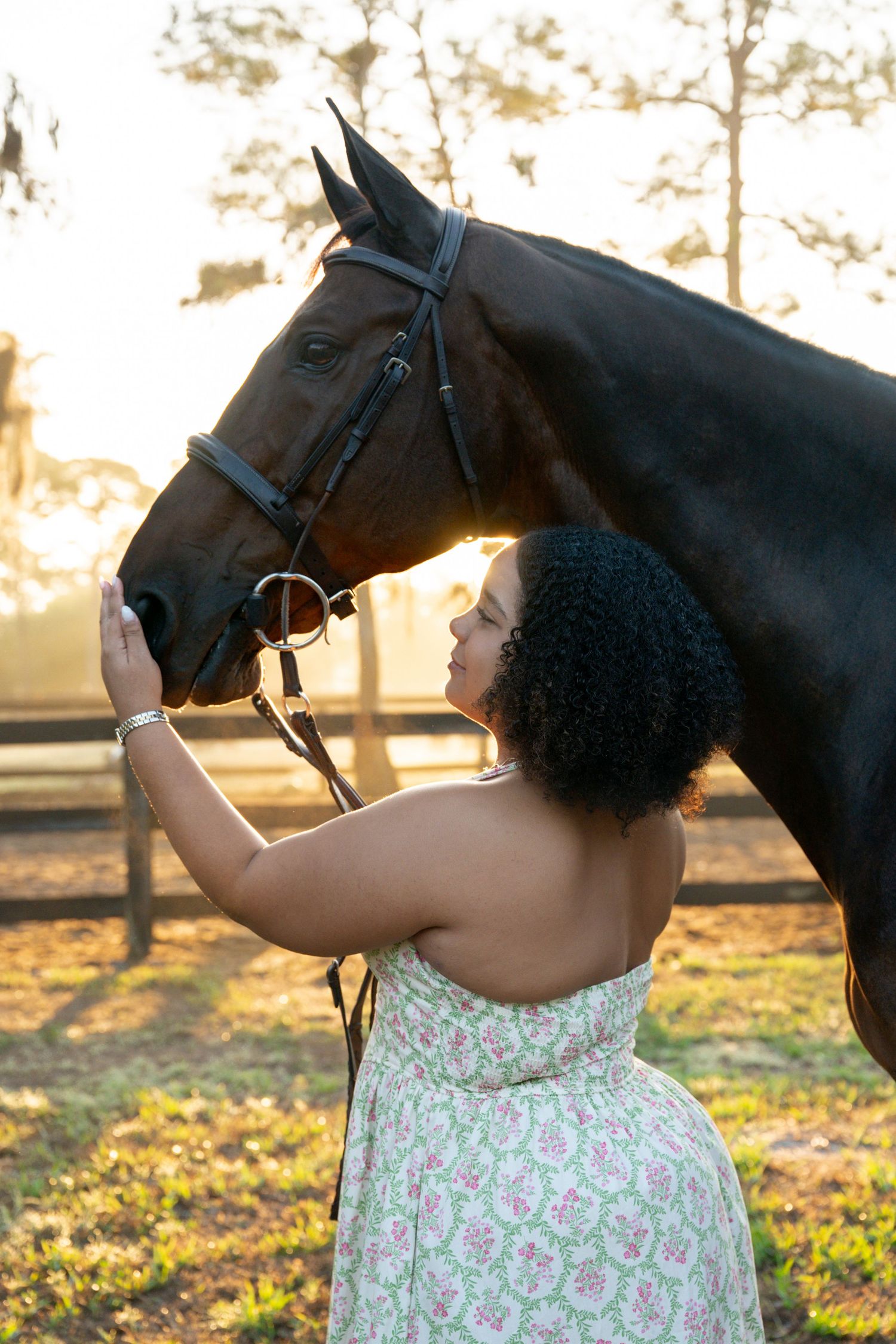 Woman in floral sundress gently holds and faces a dark bay horse at golden hour outdoors.