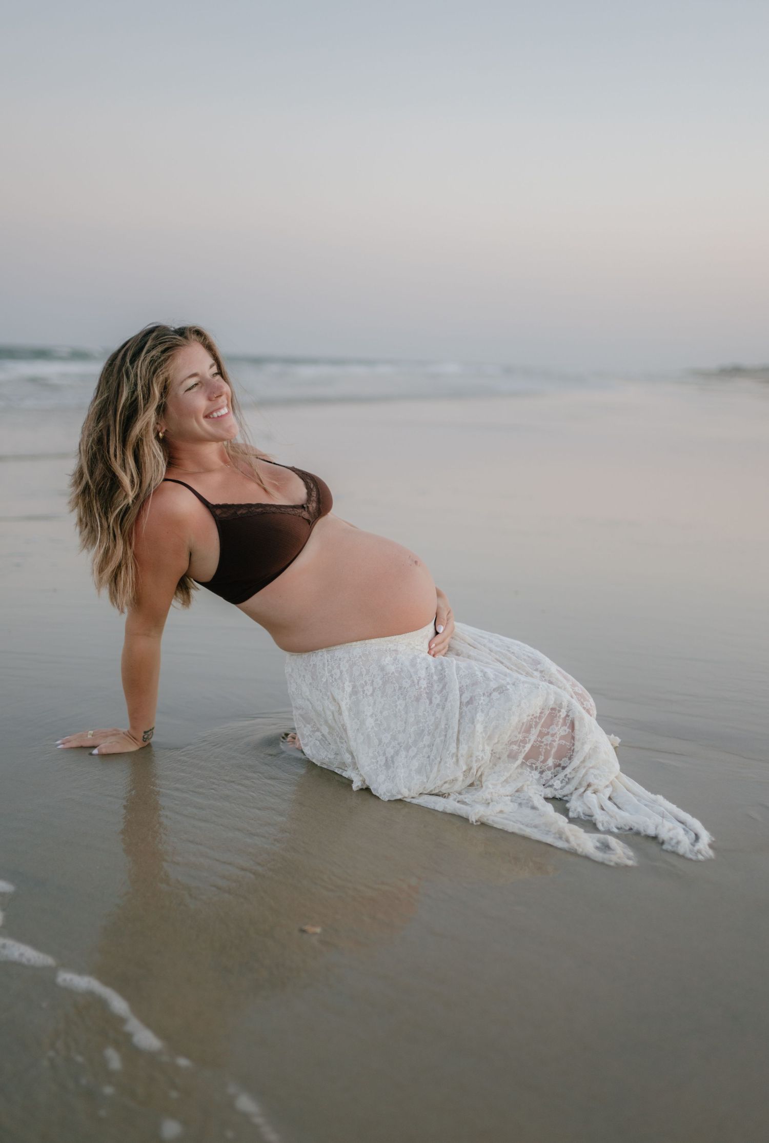 Smiling pregnant woman in black bralette and white lace skirt posing on wet beach.