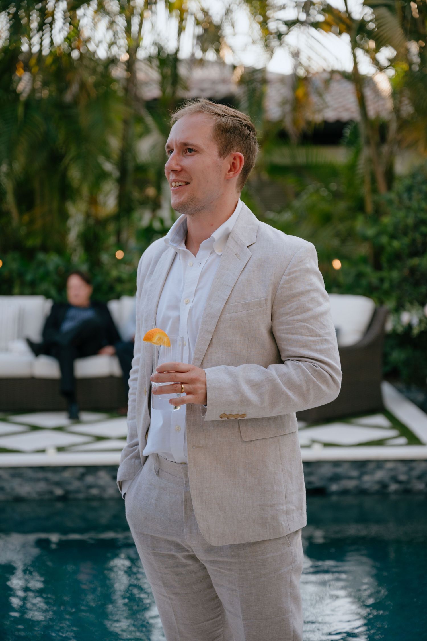 Smiling groom in a beige linen suit holding a cocktail beside a pool at an outdoor wedding reception.