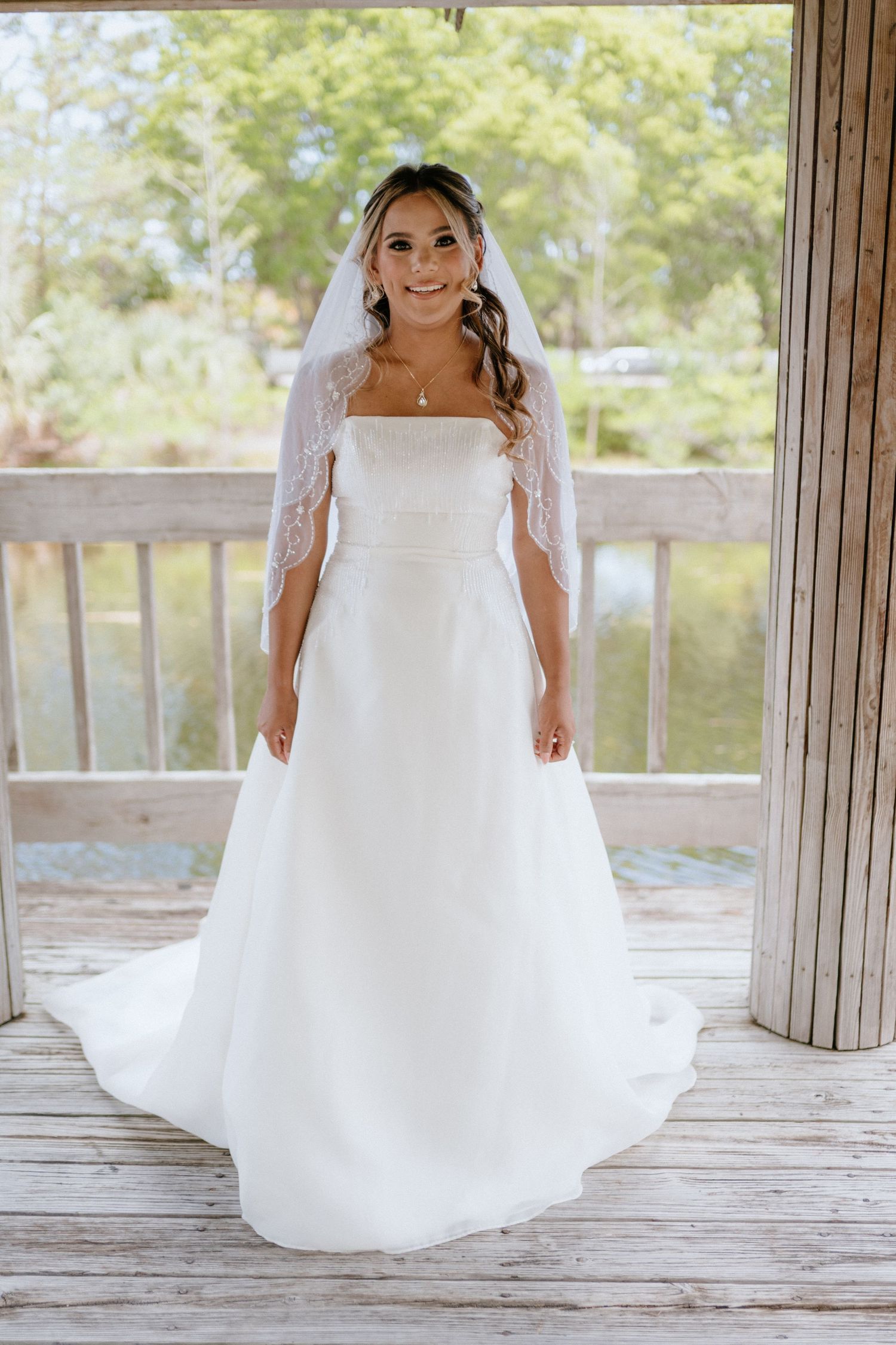 Radiant bride poses in a white A-line gown with lace veil on a wooden deck.