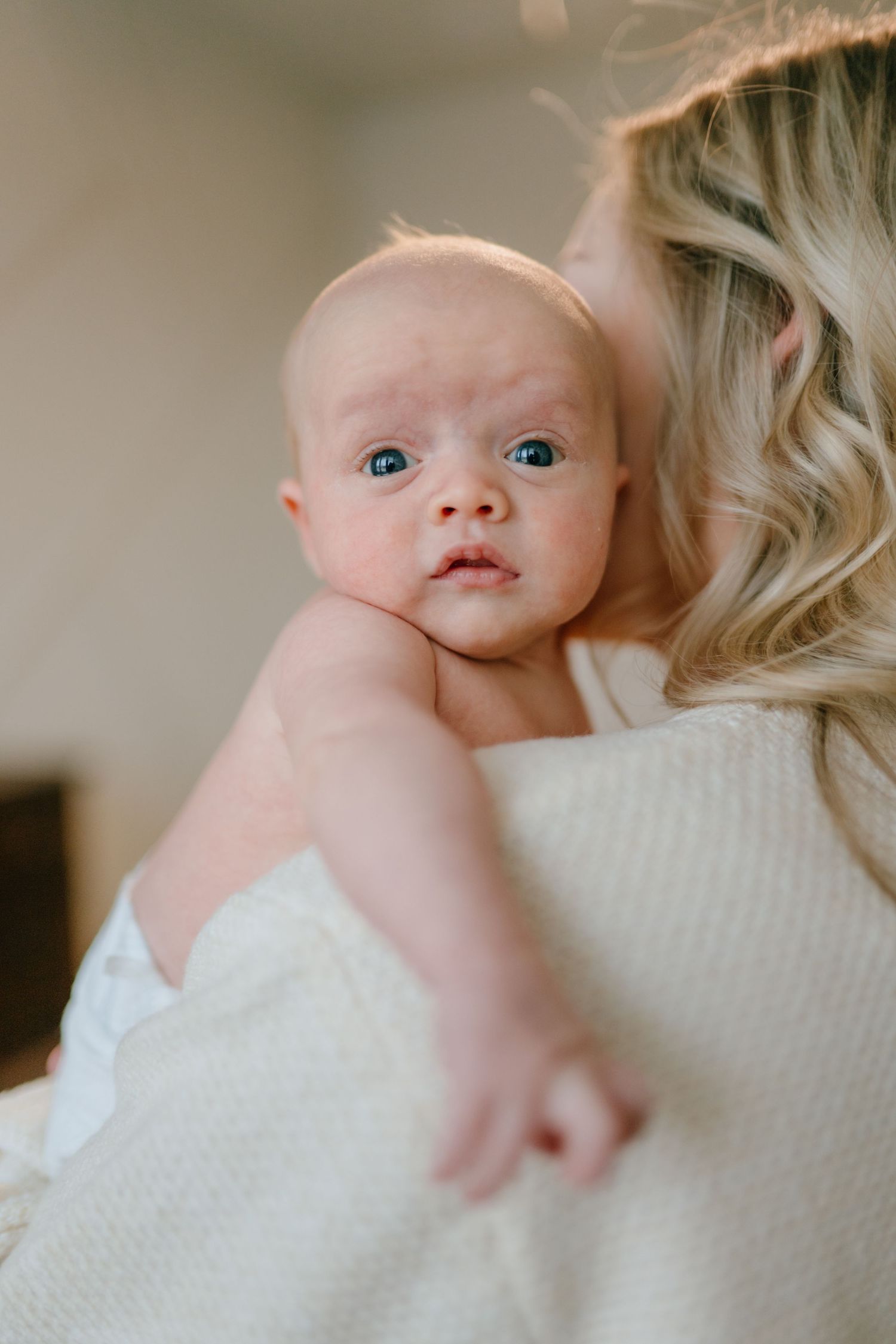 A wide-eyed newborn baby with blue eyes being held over a blonde mother's shoulder.