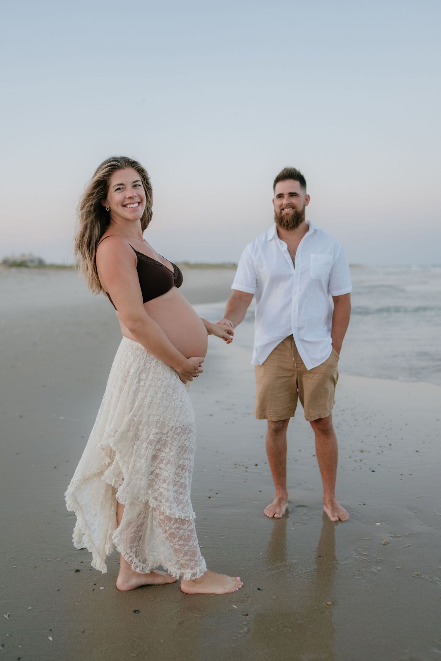 Pregnant couple holding hands on beach shoreline at dusk during maternity photoshoot.