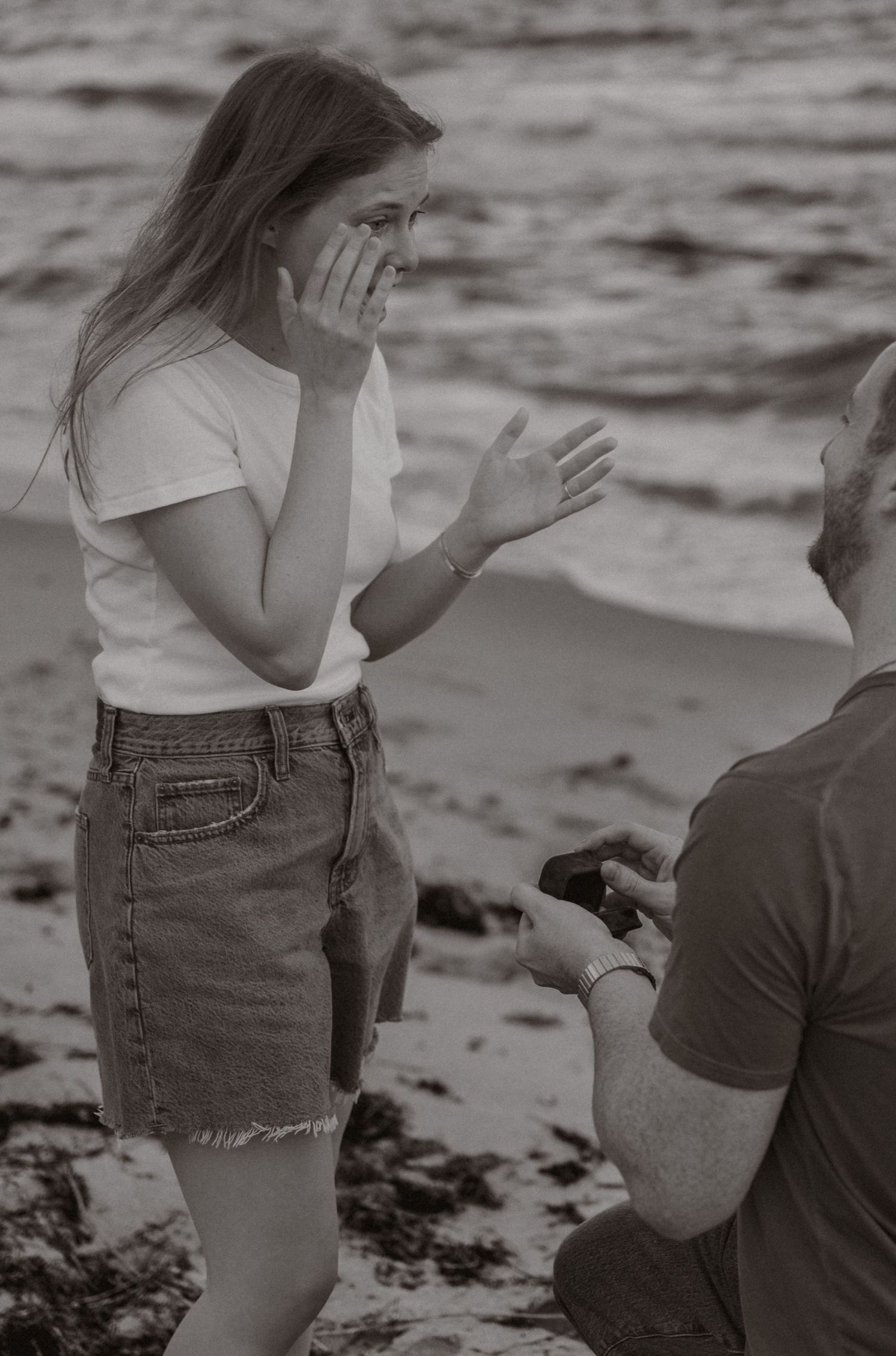 Woman reacting with joy to beach proposal, hands on face, black and white photography.