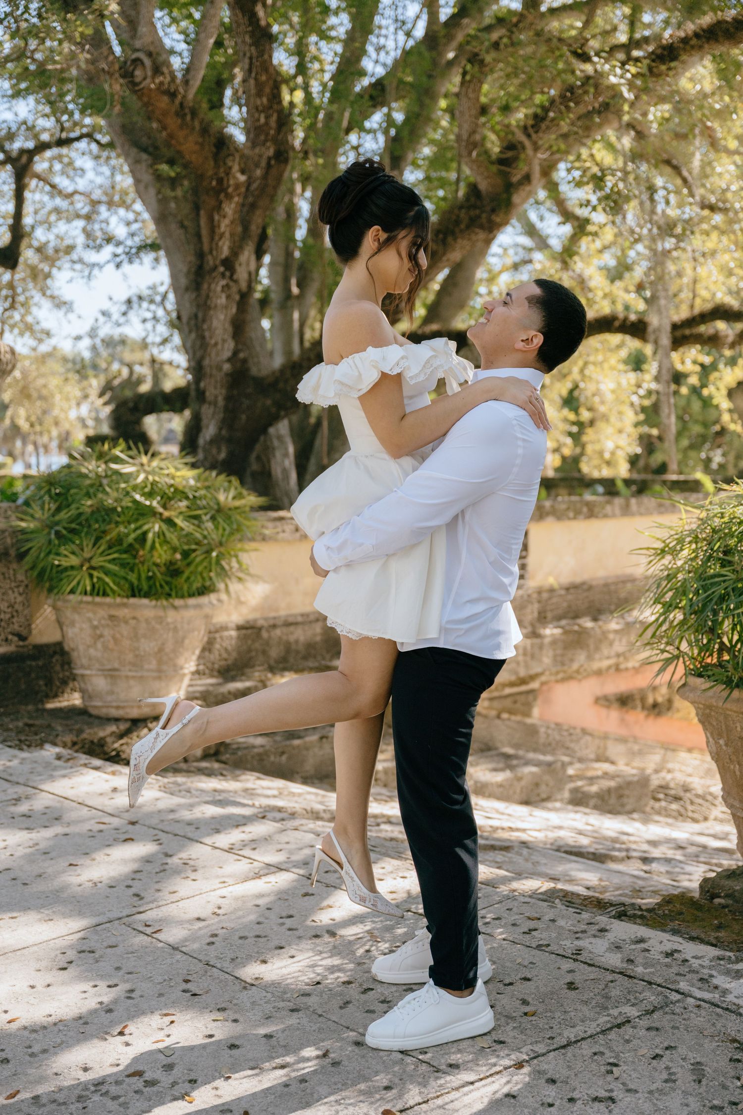Groom lifts bride in white mini dress in a romantic garden setting with lush trees.