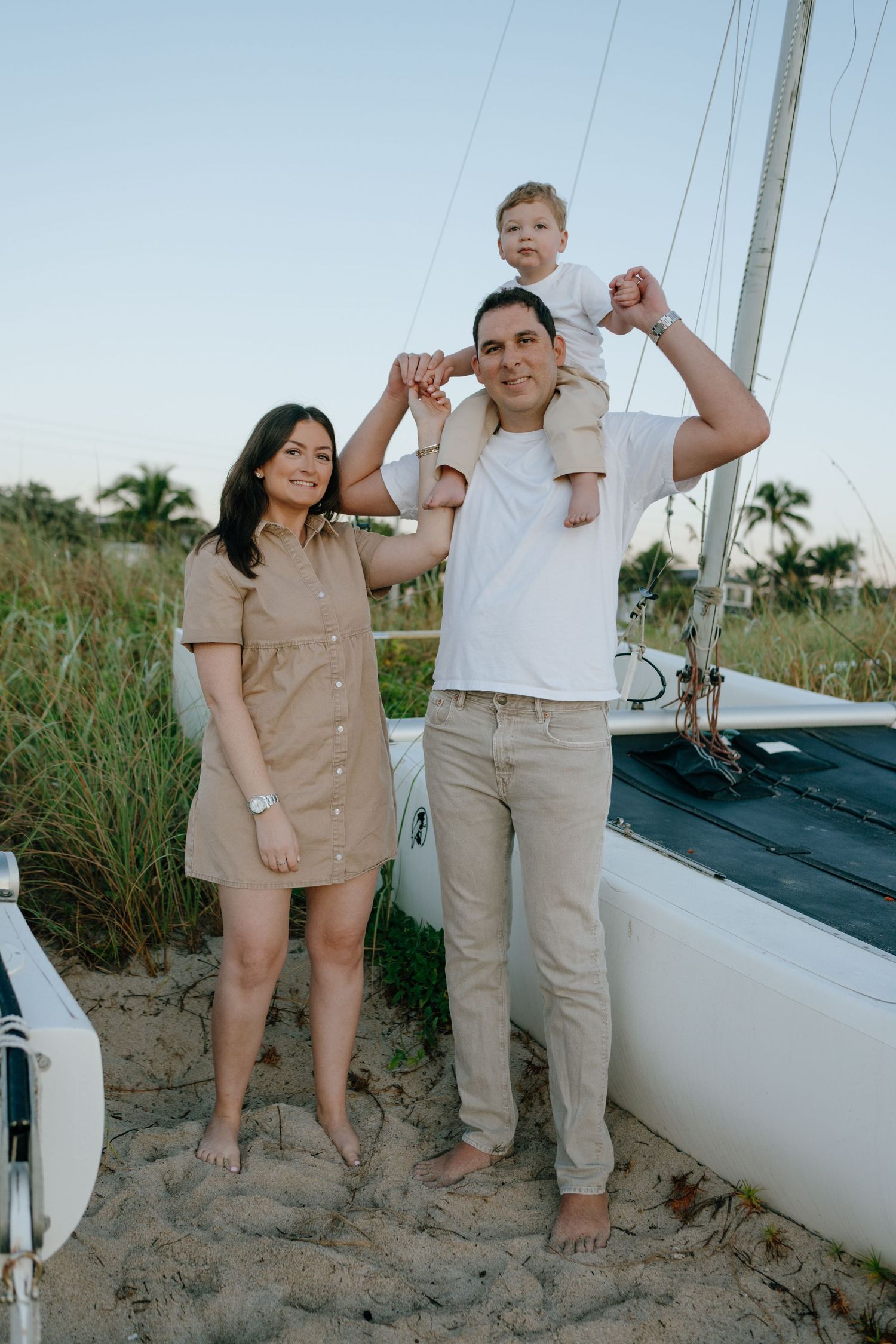 Smiling family with toddler on dad's shoulders standing next to a sailboat on a sandy beach at sunset.
