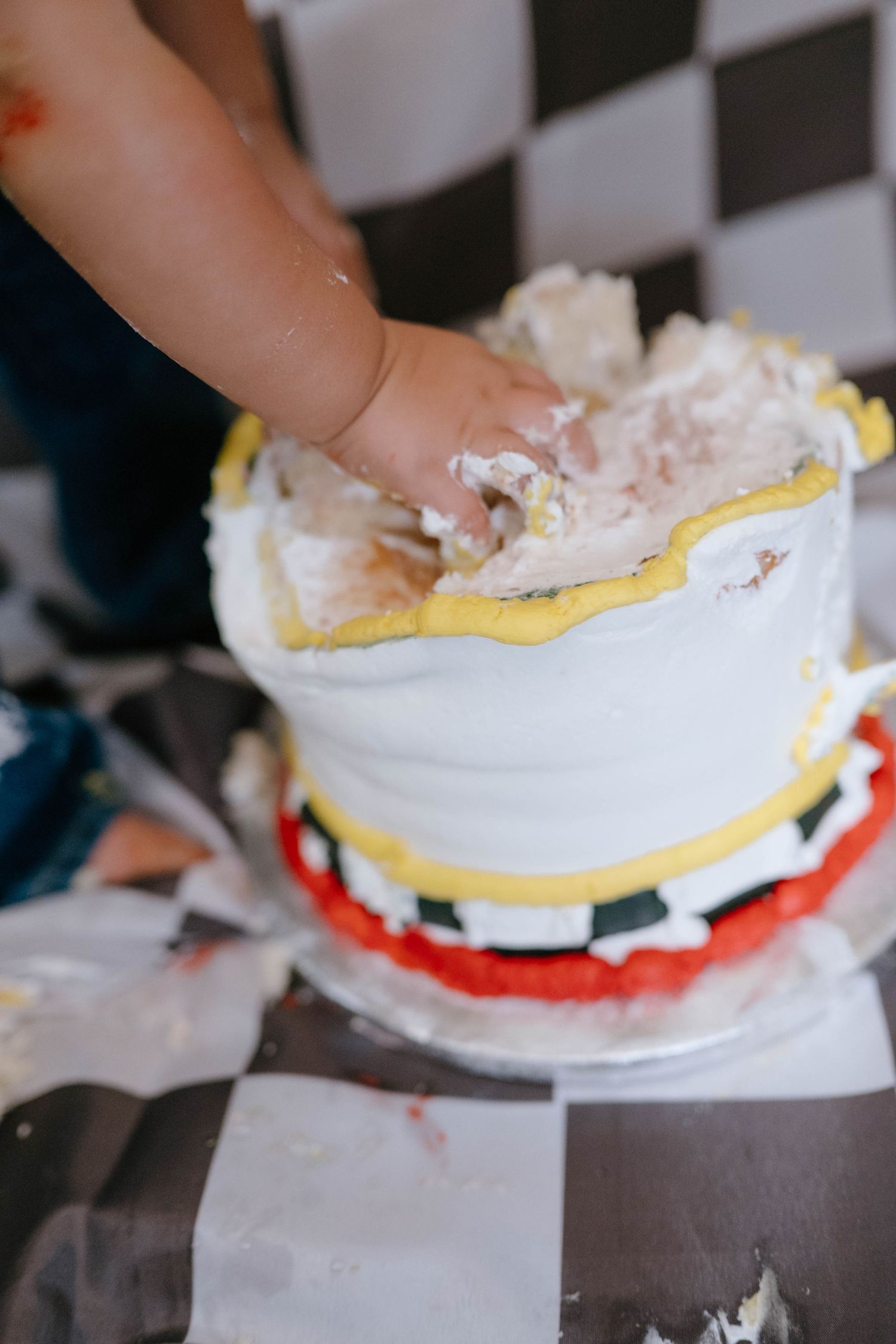 Baby hand smashing into a white and yellow first birthday cake on a checkered surface.