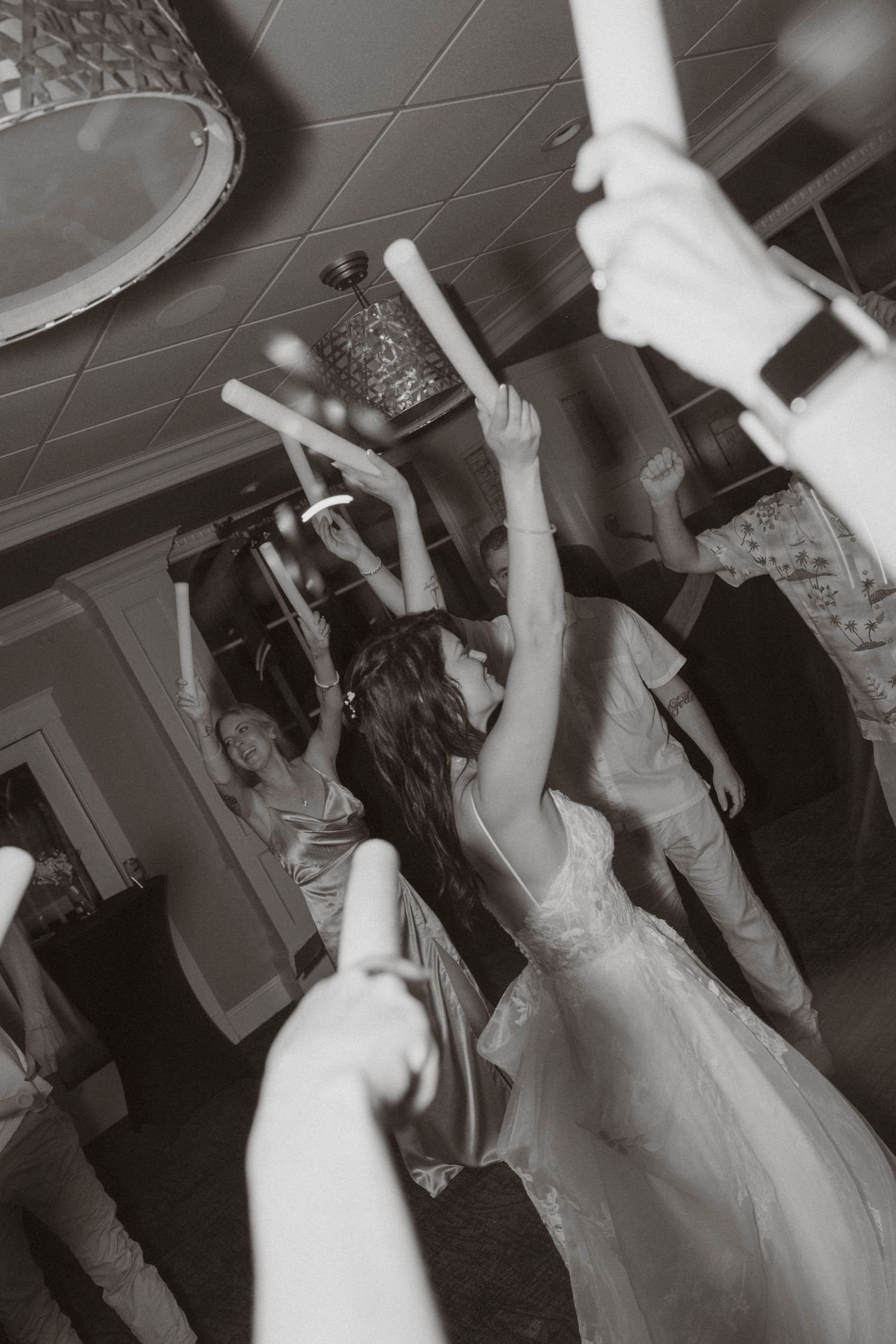 Black and white photo of wedding guests dancing and raising glow sticks at a lively reception party.