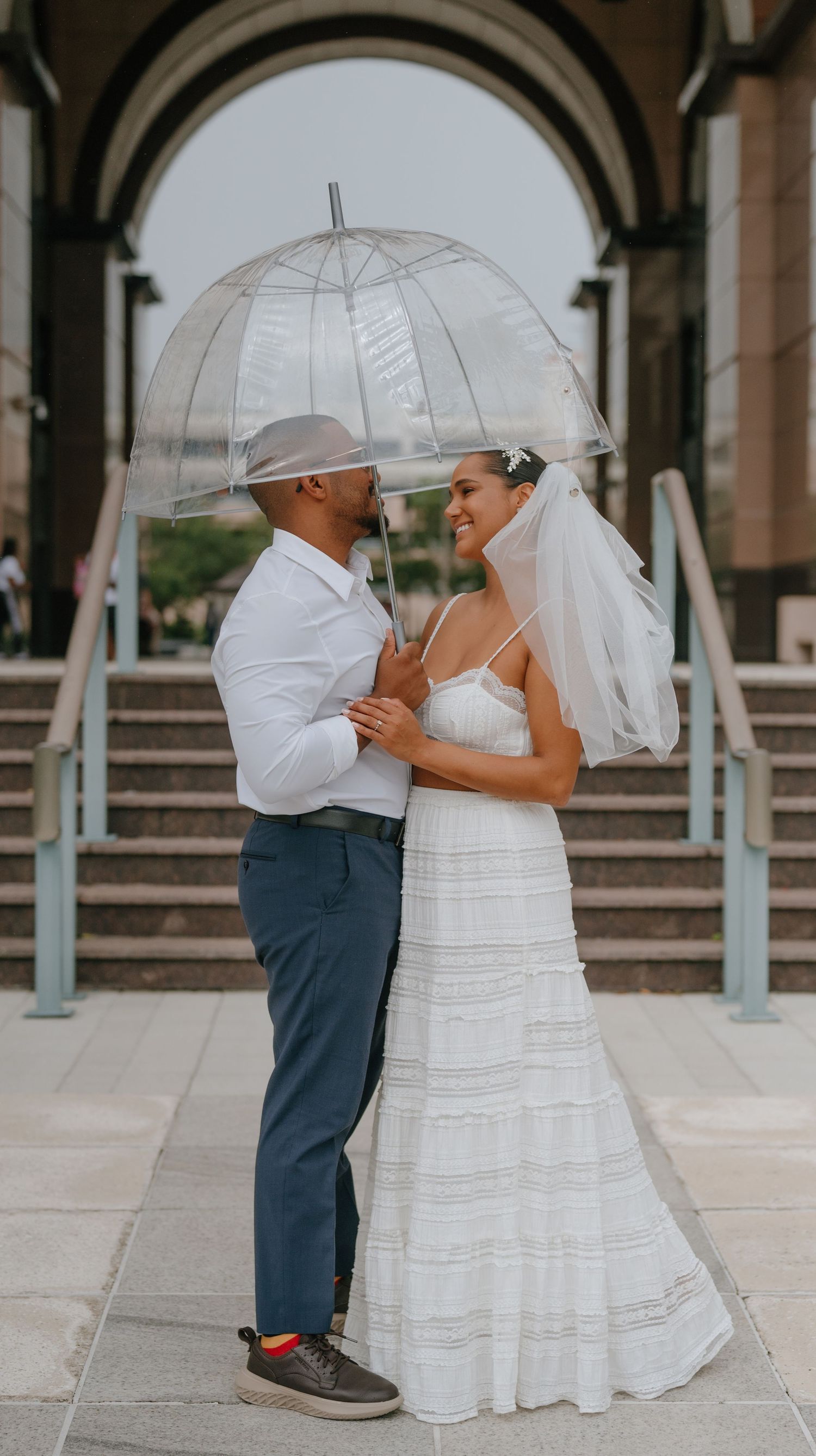 Bride and groom standing under a clear bubble umbrella outside an arched building on their wedding day.