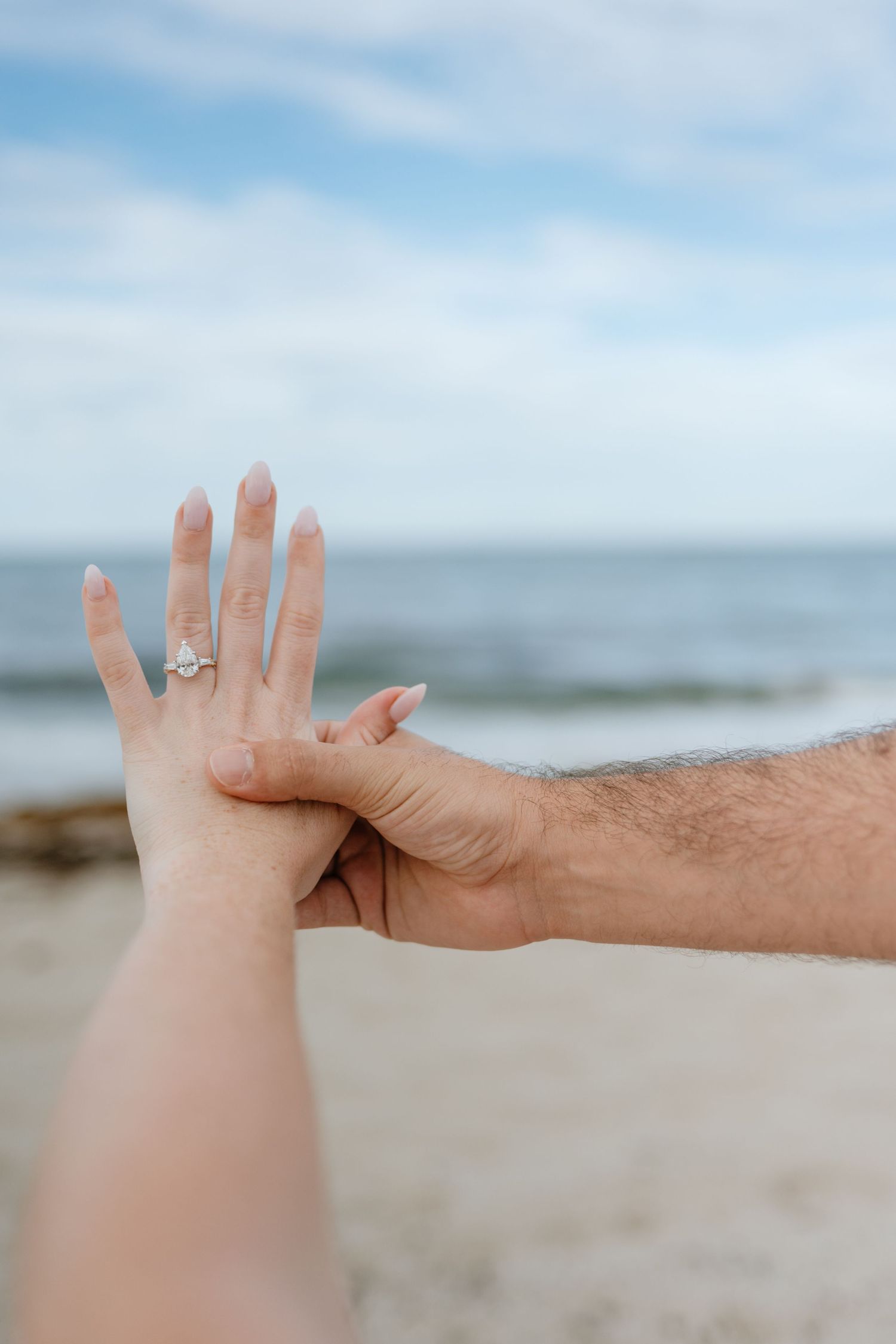 Close-up of man holding woman's hand displaying a sparkling diamond engagement ring on the beach.
