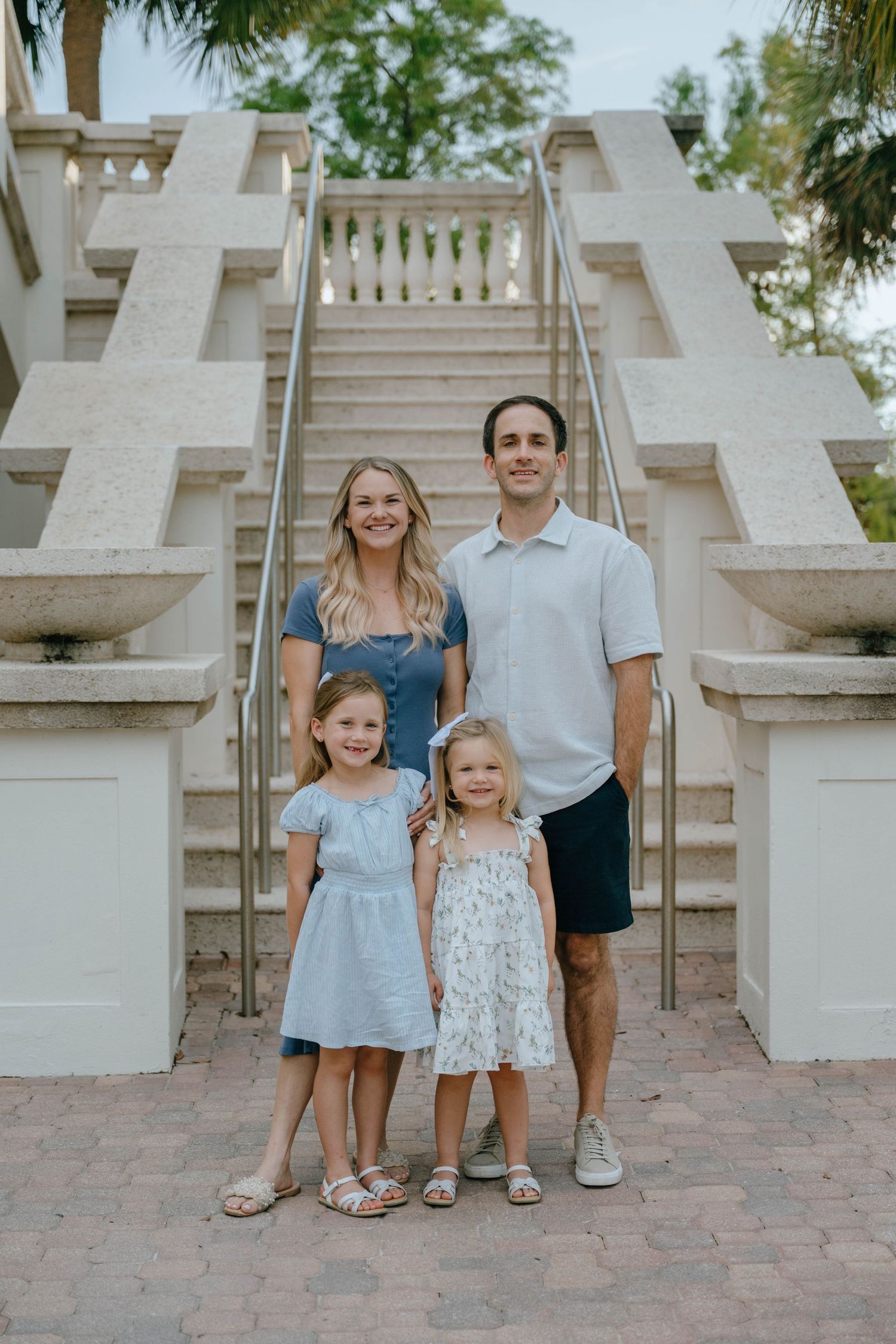 A family of four smiles together in front of an elegant outdoor staircase at dusk.
