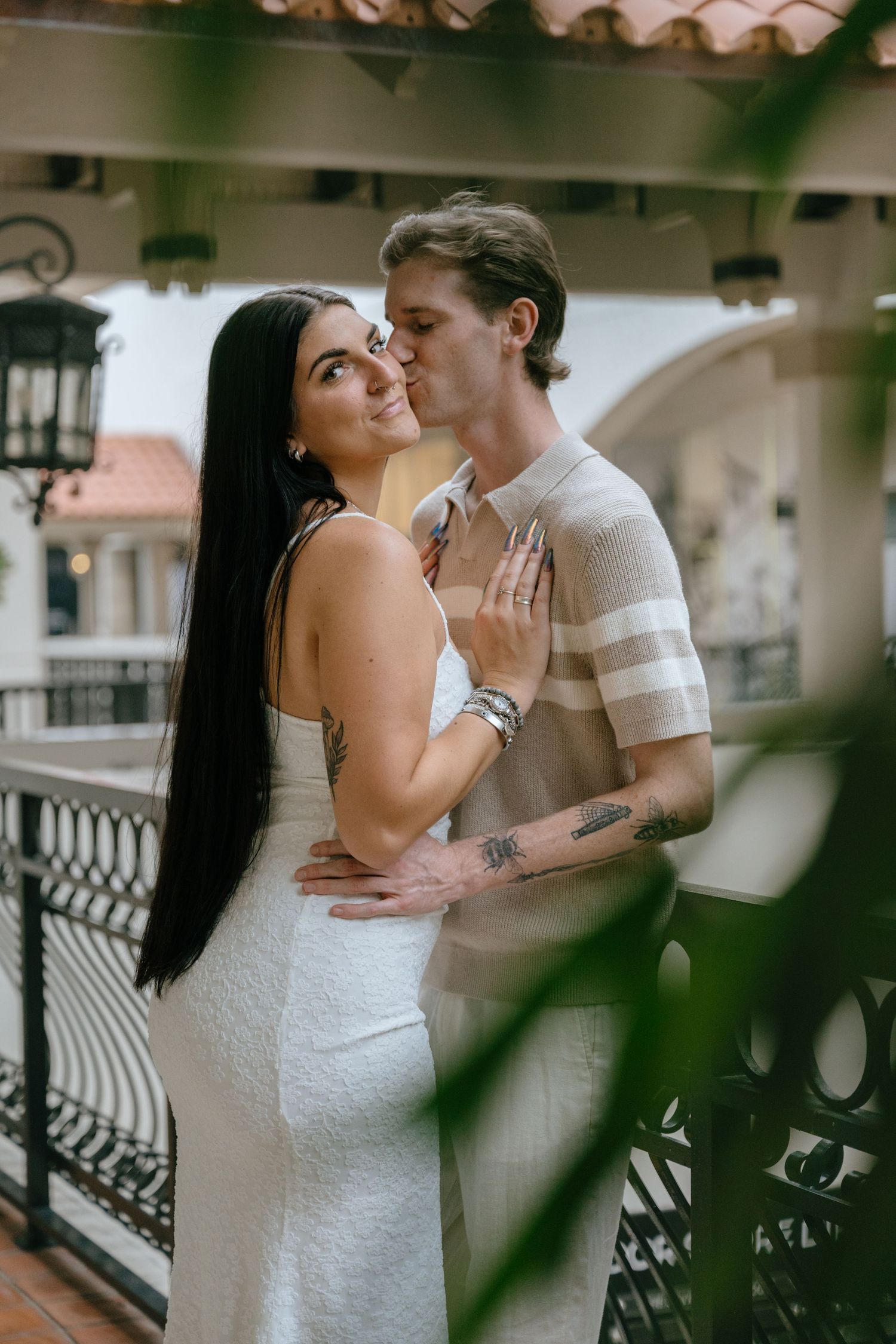 Tattooed couple sharing a sweet kiss on a decorative balcony, framed by lush greenery in Palm Beach.