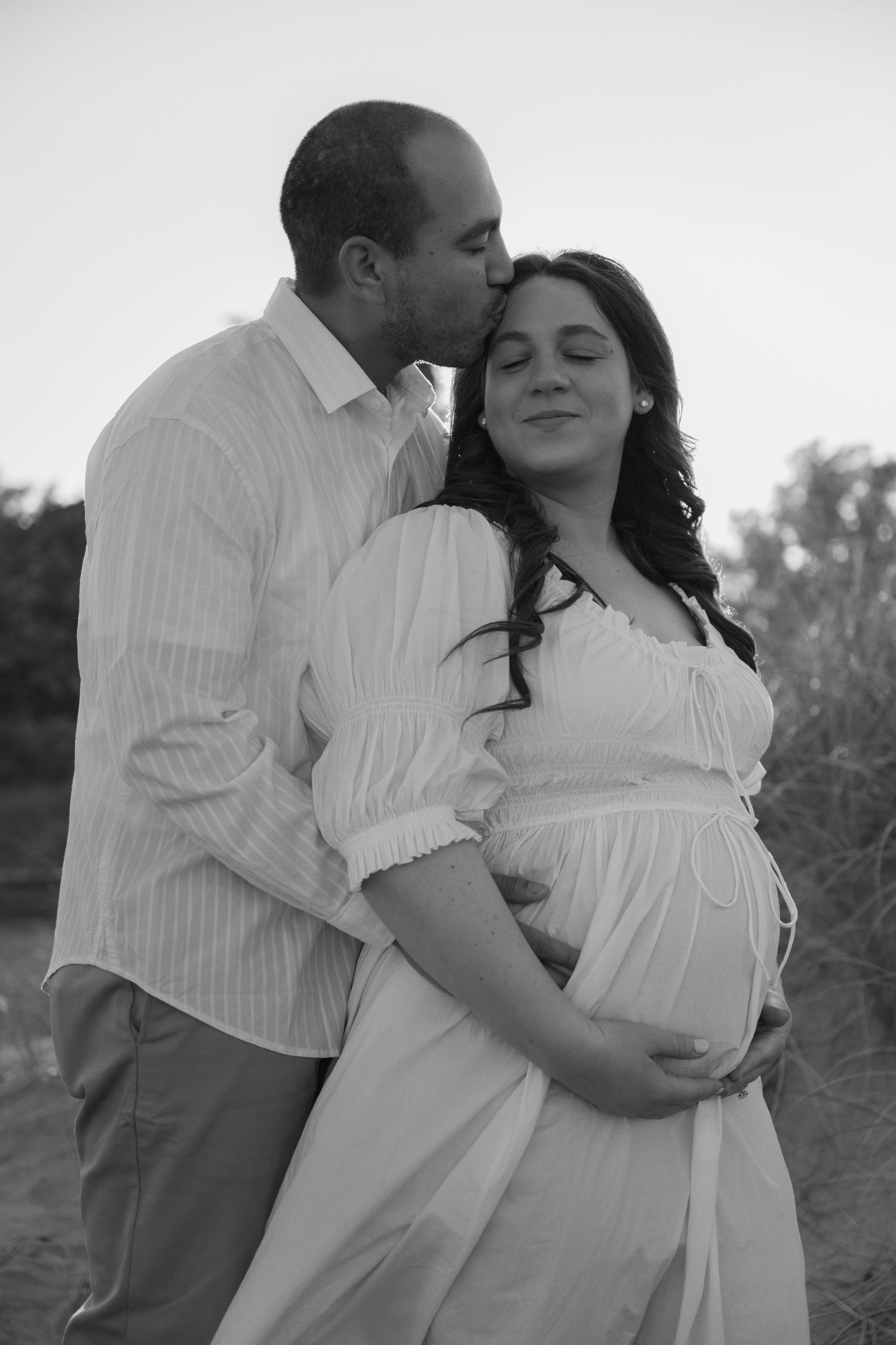 Black and white photo of man kissing pregnant wife on forehead outdoors at dusk.