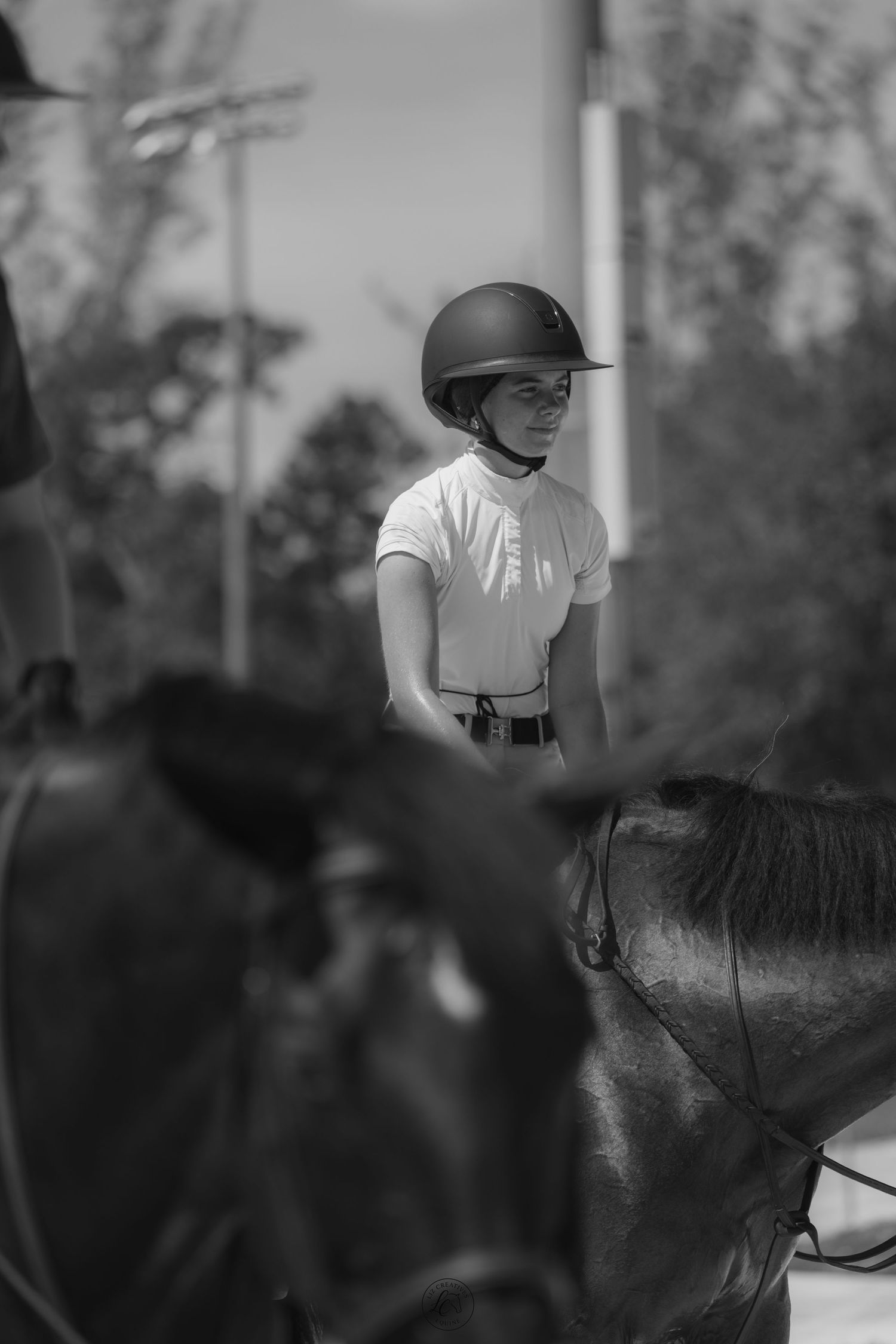 Black and white portrait of young equestrian rider in helmet and white shirt seated on dark horse outdoors.