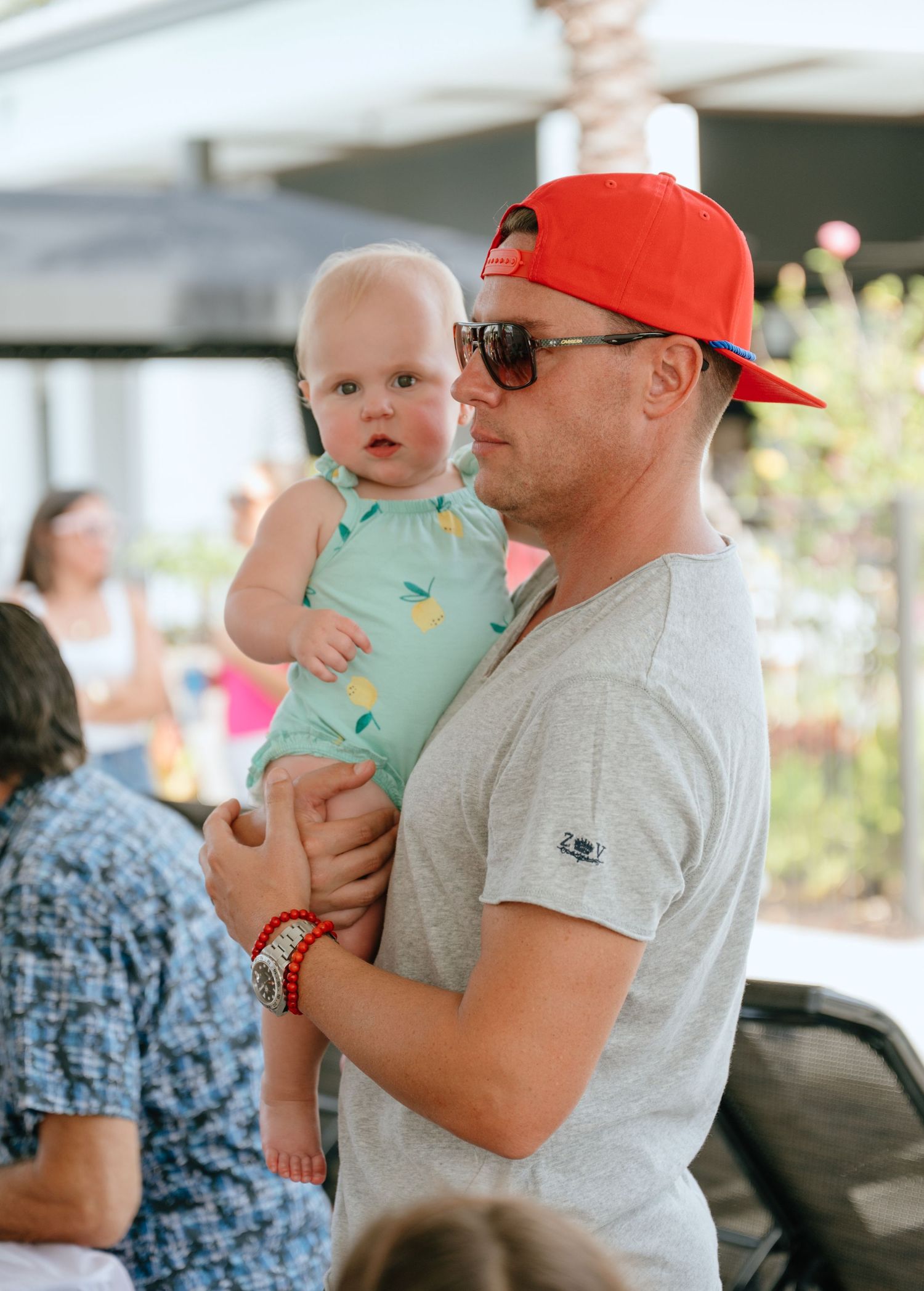 Father in red cap and sunglasses tenderly holds his baby girl in a mint green outfit outdoors.