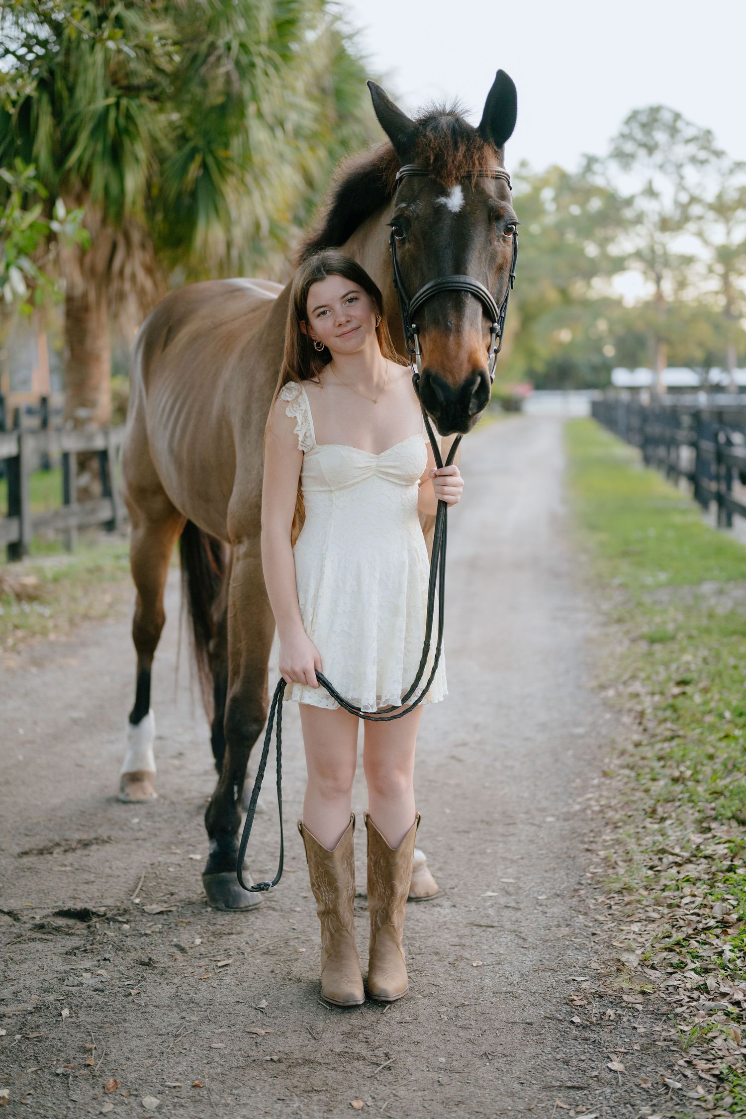 Young woman in white dress and cowboy boots stands proudly with a bay horse on a farm path.