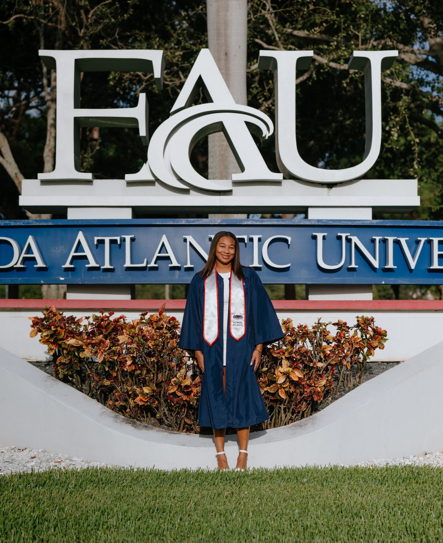 Graduate in navy cap and gown posing in front of Florida Atlantic University sign.