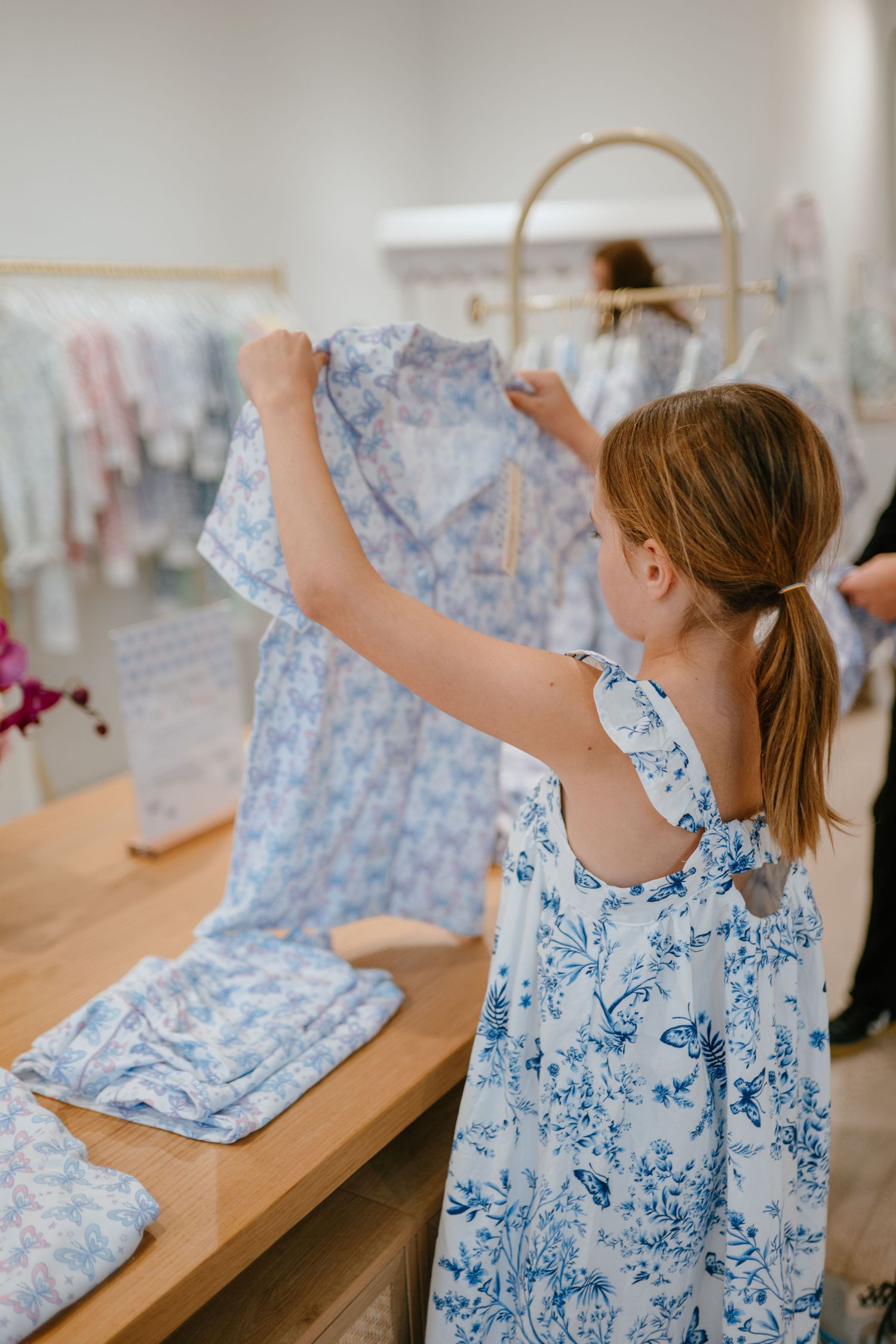 Young girl in floral dress examining blue butterfly print clothing at a boutique store wooden display table.