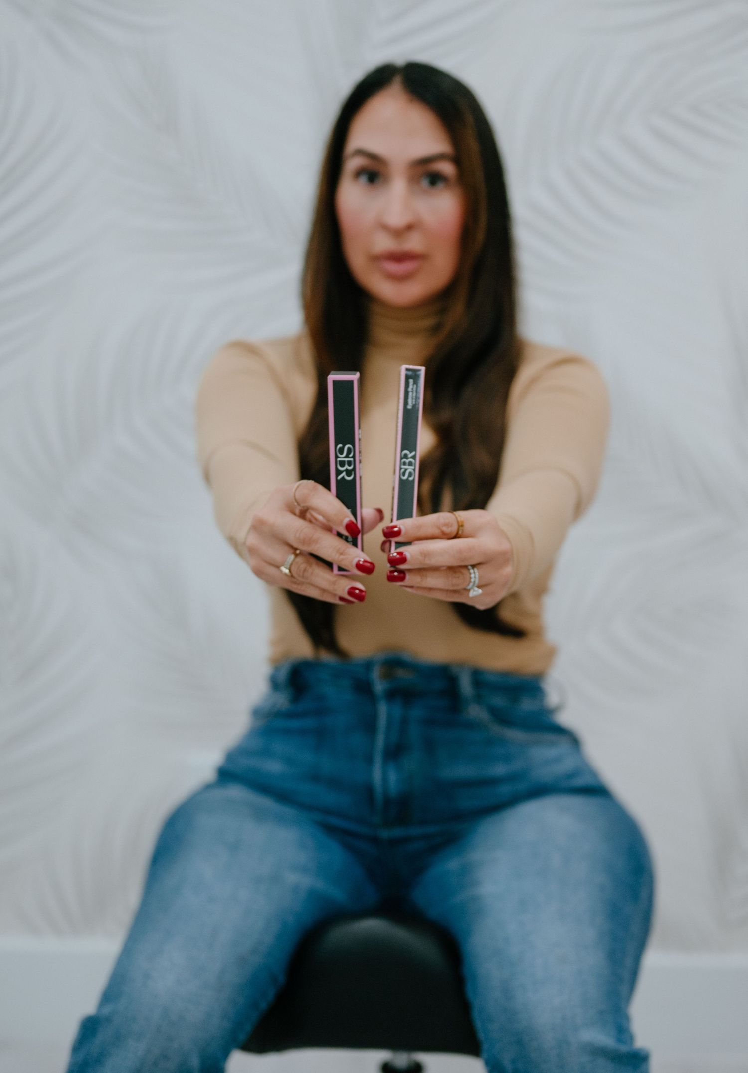 Woman holding two KVD beauty products toward camera, wearing beige turtleneck and jeans with red manicure.
