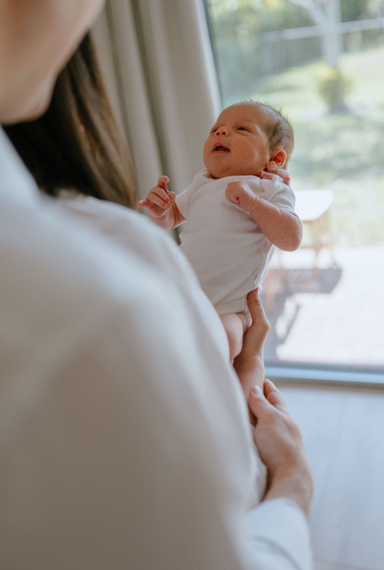 Mother gently lifts her newborn baby near a bright window in a white room.