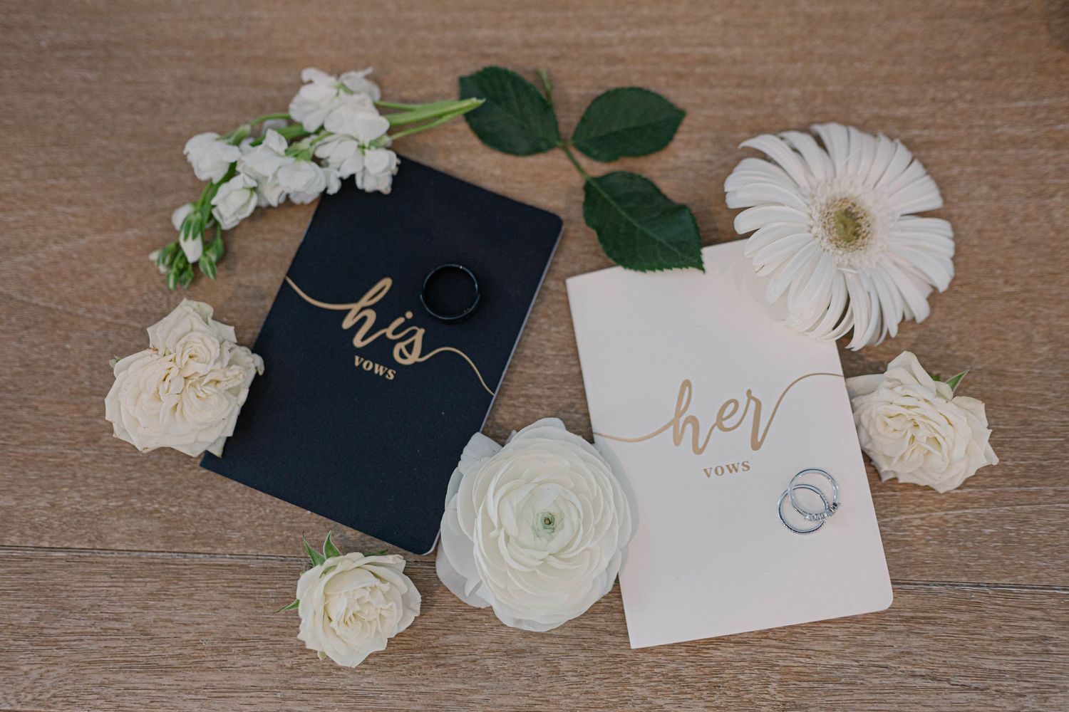 His and hers wedding vow books with gold lettering surrounded by white roses and flowers.