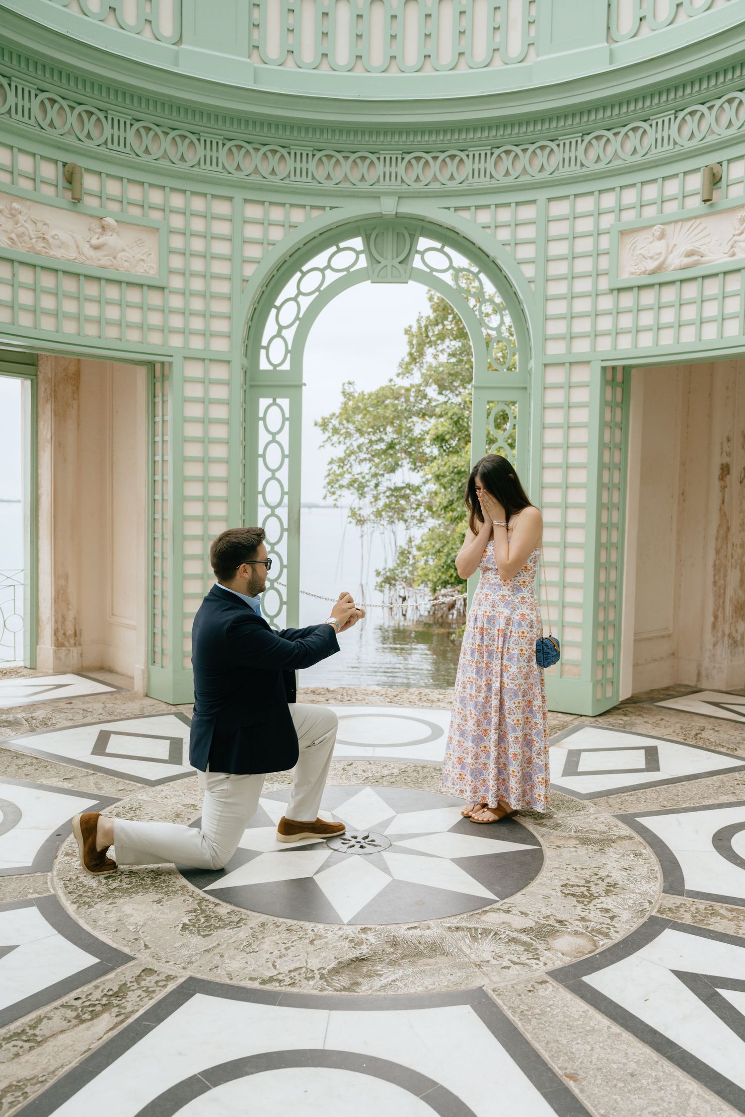 Man proposing on one knee to surprised woman in floral dress inside ornate green pavilion interior.