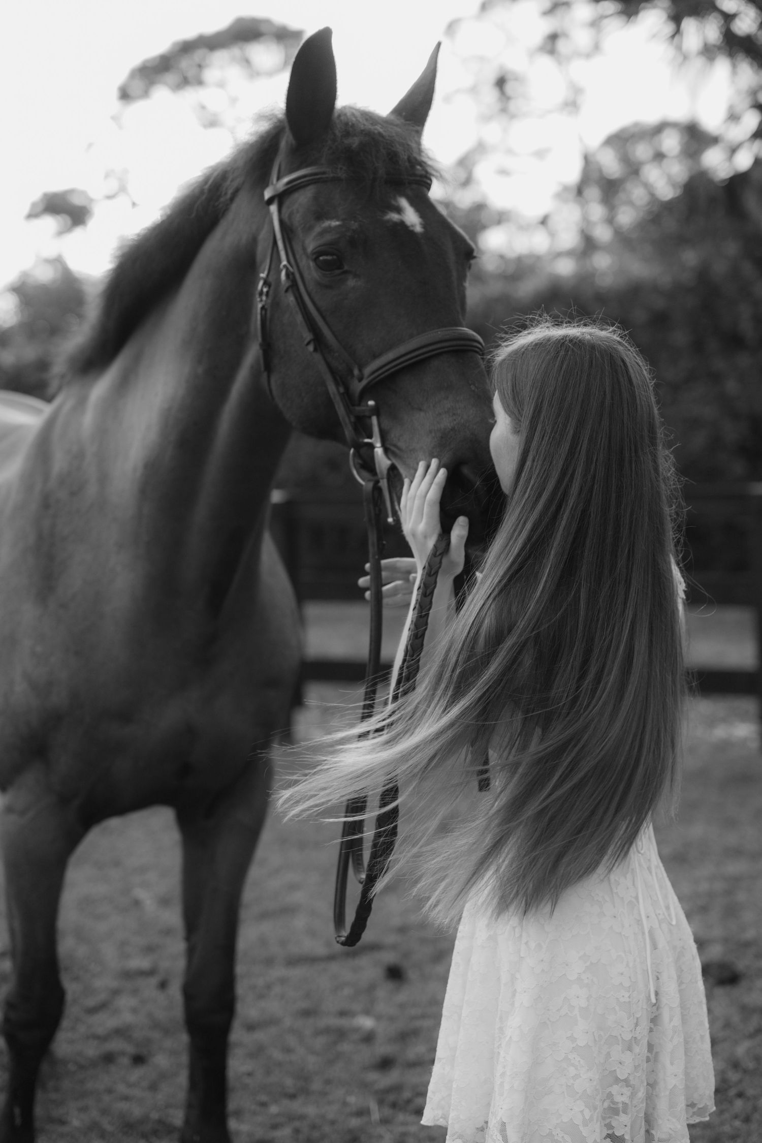 Black and white photo of a long-haired girl gently kissing a bridled bay horse outdoors.