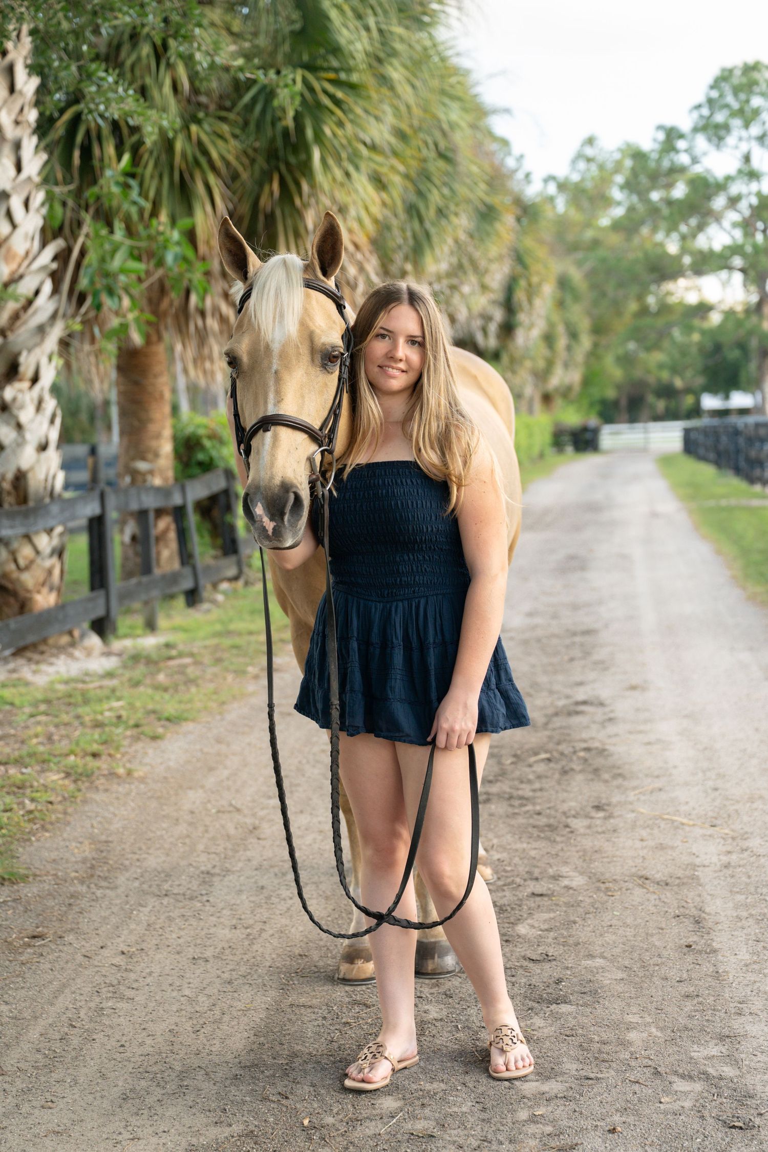 Smiling young woman in navy dress poses with palomino horse on palm-lined gravel path at horse stable.