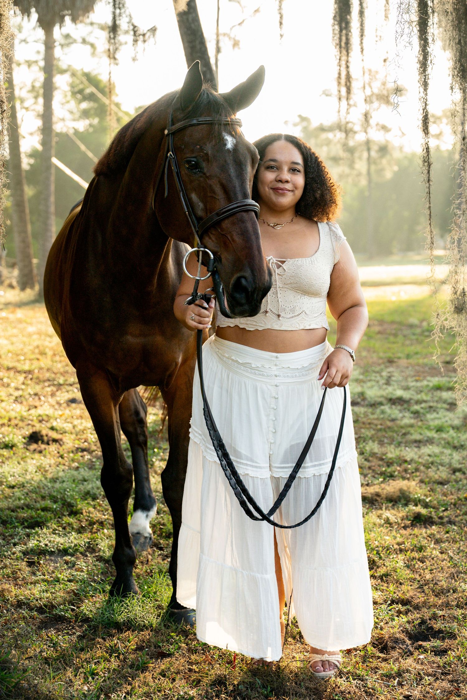 Young woman in white crop top and skirt stands with dark bay horse in sunlit mossy forest.