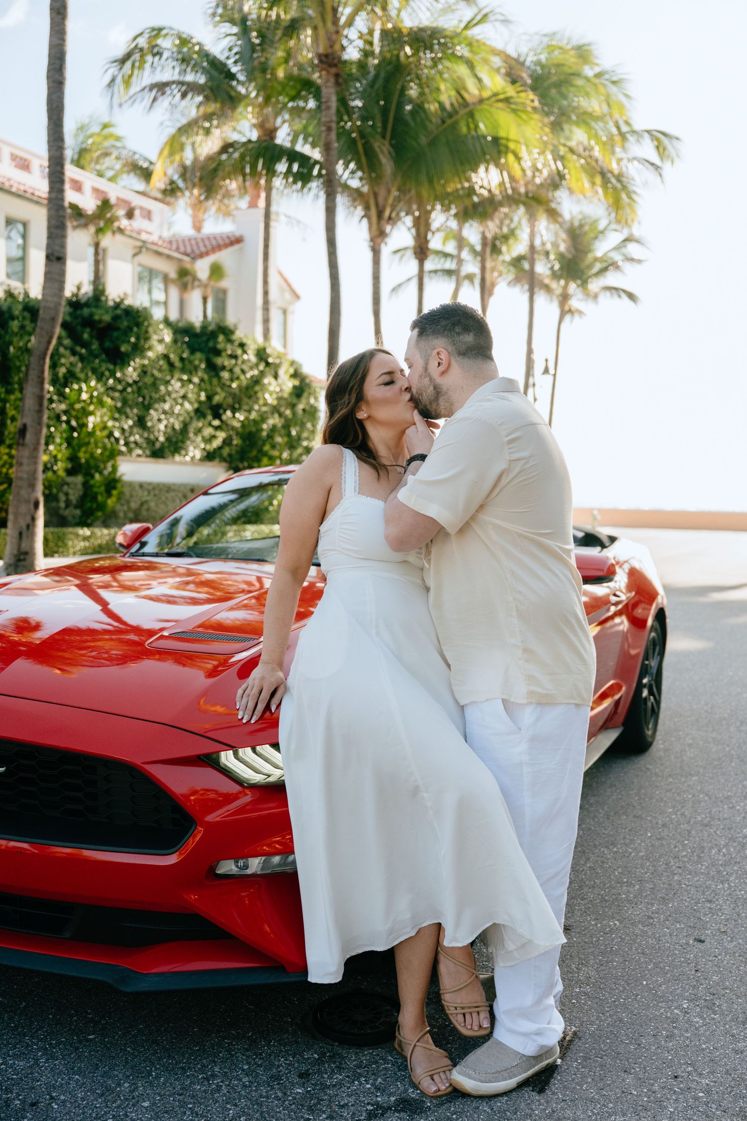 Couple sharing a romantic kiss leaning against a red Ford Mustang convertible surrounded by tropical palm trees.