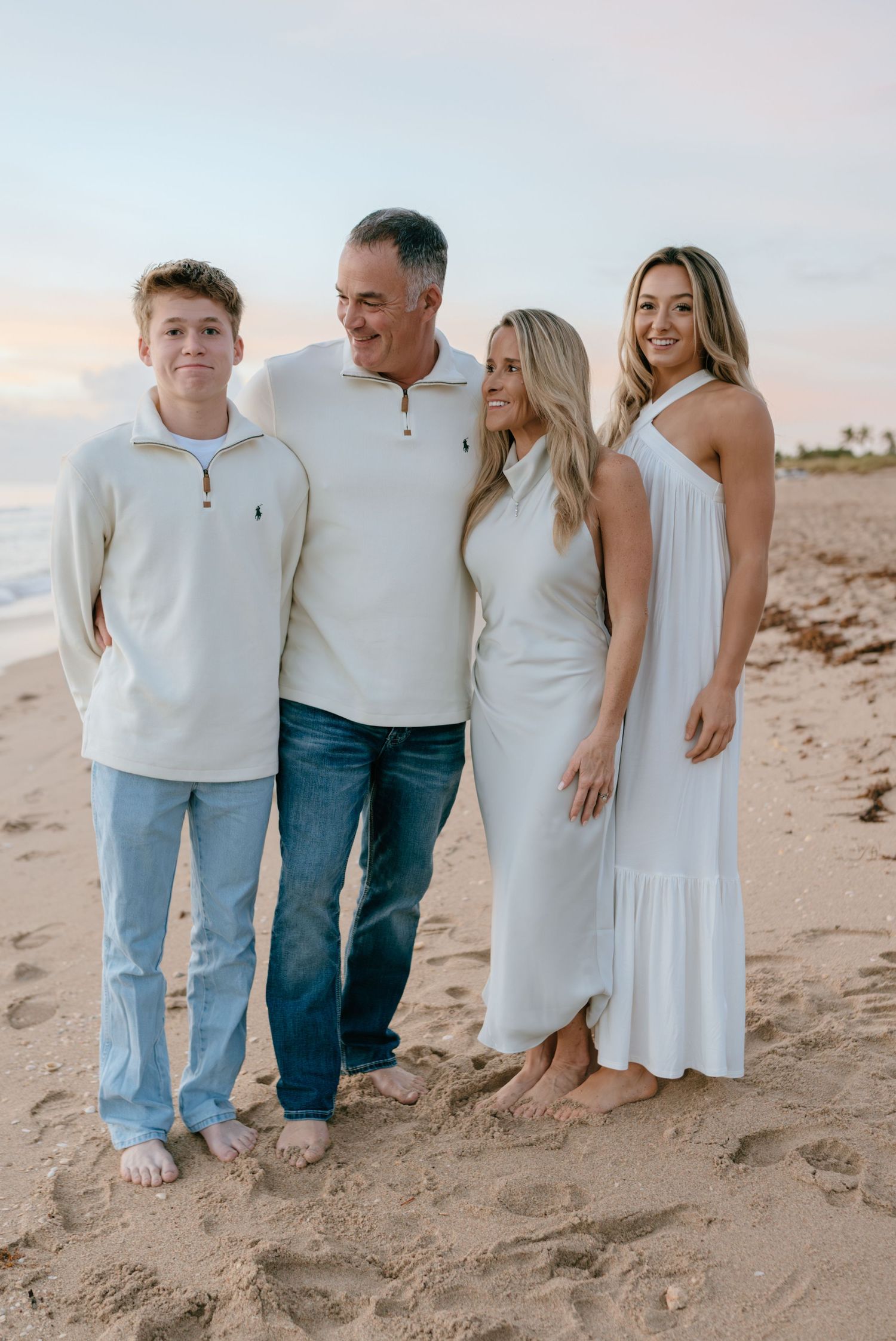 A smiling family of four stands together on a sandy beach at sunset.