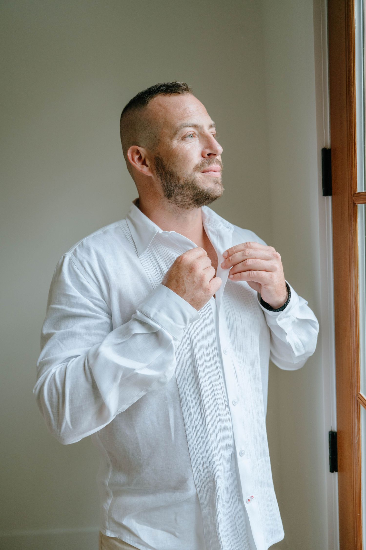 Groom in white linen shirt buttoning up while gazing out a window before his wedding.