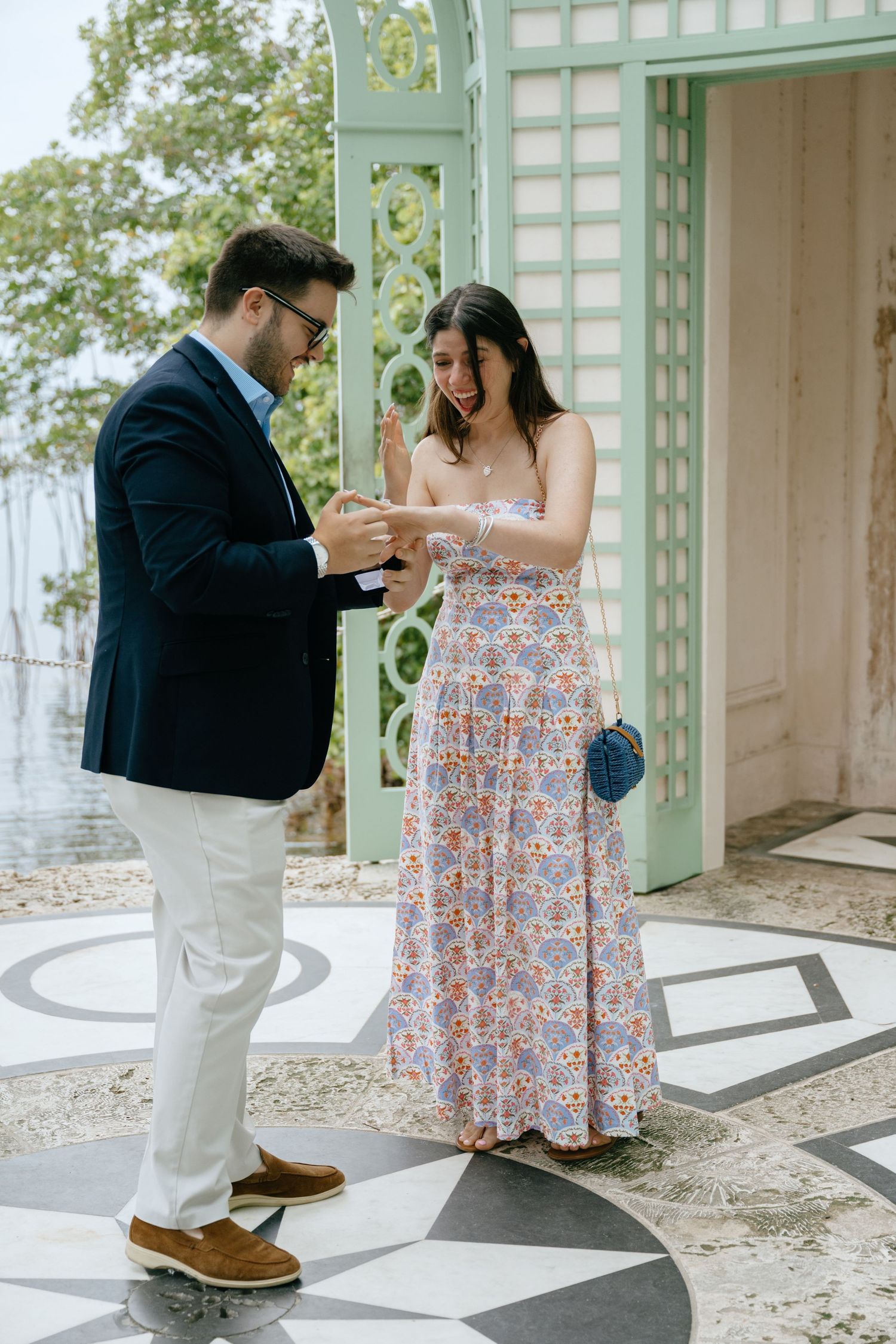 A man places an engagement ring on a woman's finger outdoors near ornate green gates.