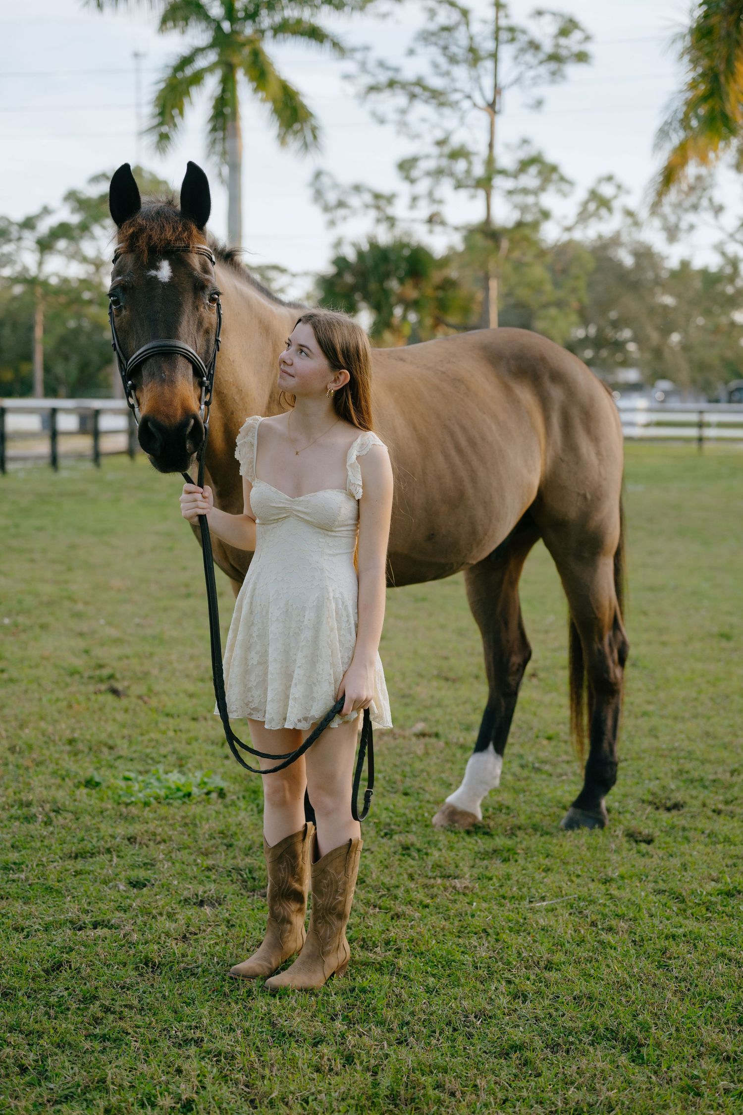 A teenage girl in a white mini dress and cowboy boots stands beside a buckskin horse on green grass.