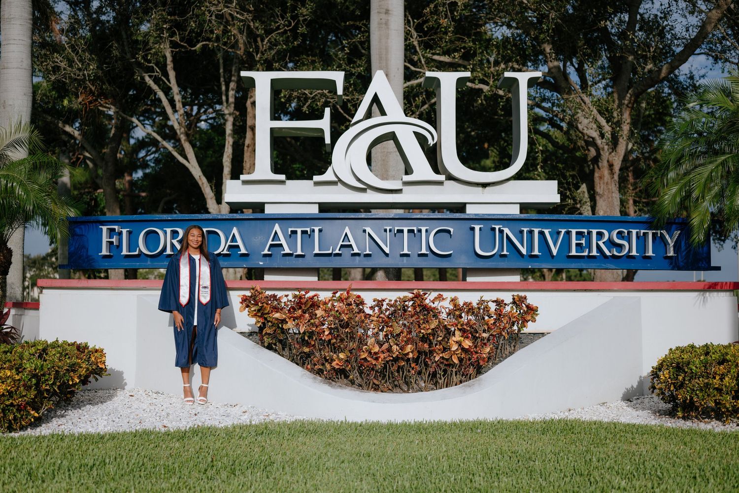 Graduate in navy cap and gown standing before Florida Atlantic University entrance sign.