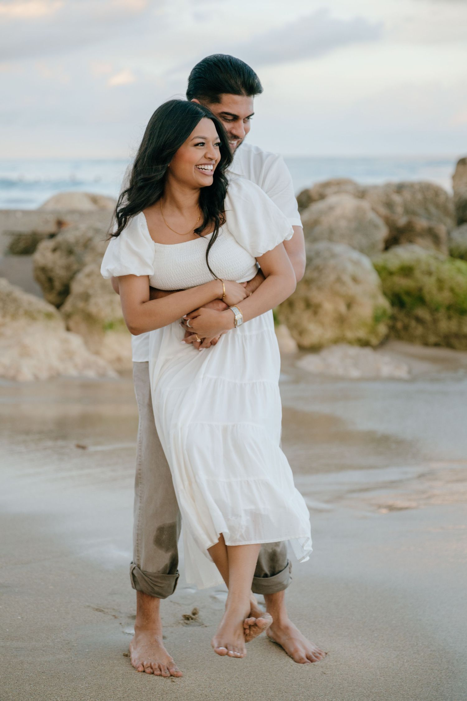 Couple embracing on a beach, woman in white dress smiling, man holding her from behind near rocky shore.