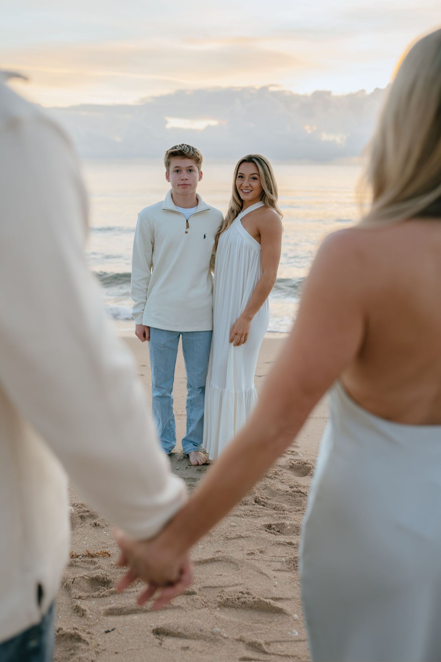 Two teenagers smile at their parents holding hands in the foreground on a beach.