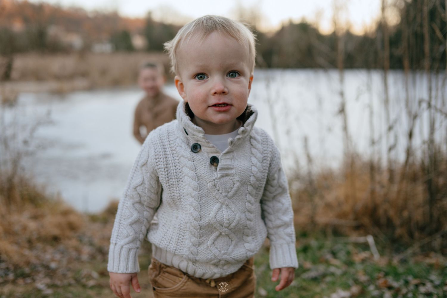 Close-up portrait of a blond toddler boy wearing a cream cable-knit sweater outdoors near a pond.