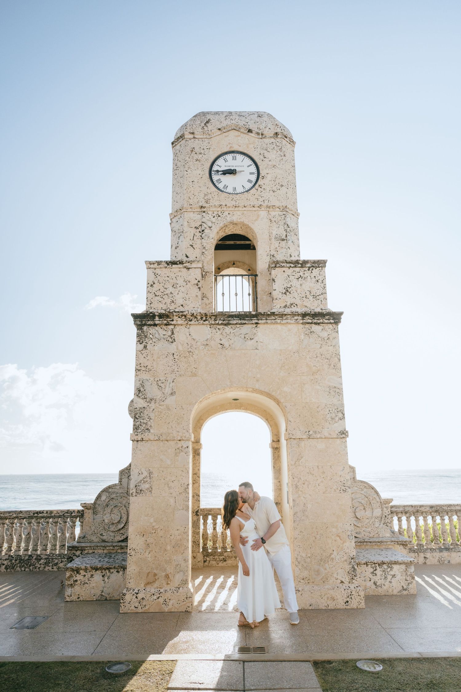 Couple sharing a tender kiss under a historic stone clock tower arch with ocean views in Palm Beach.