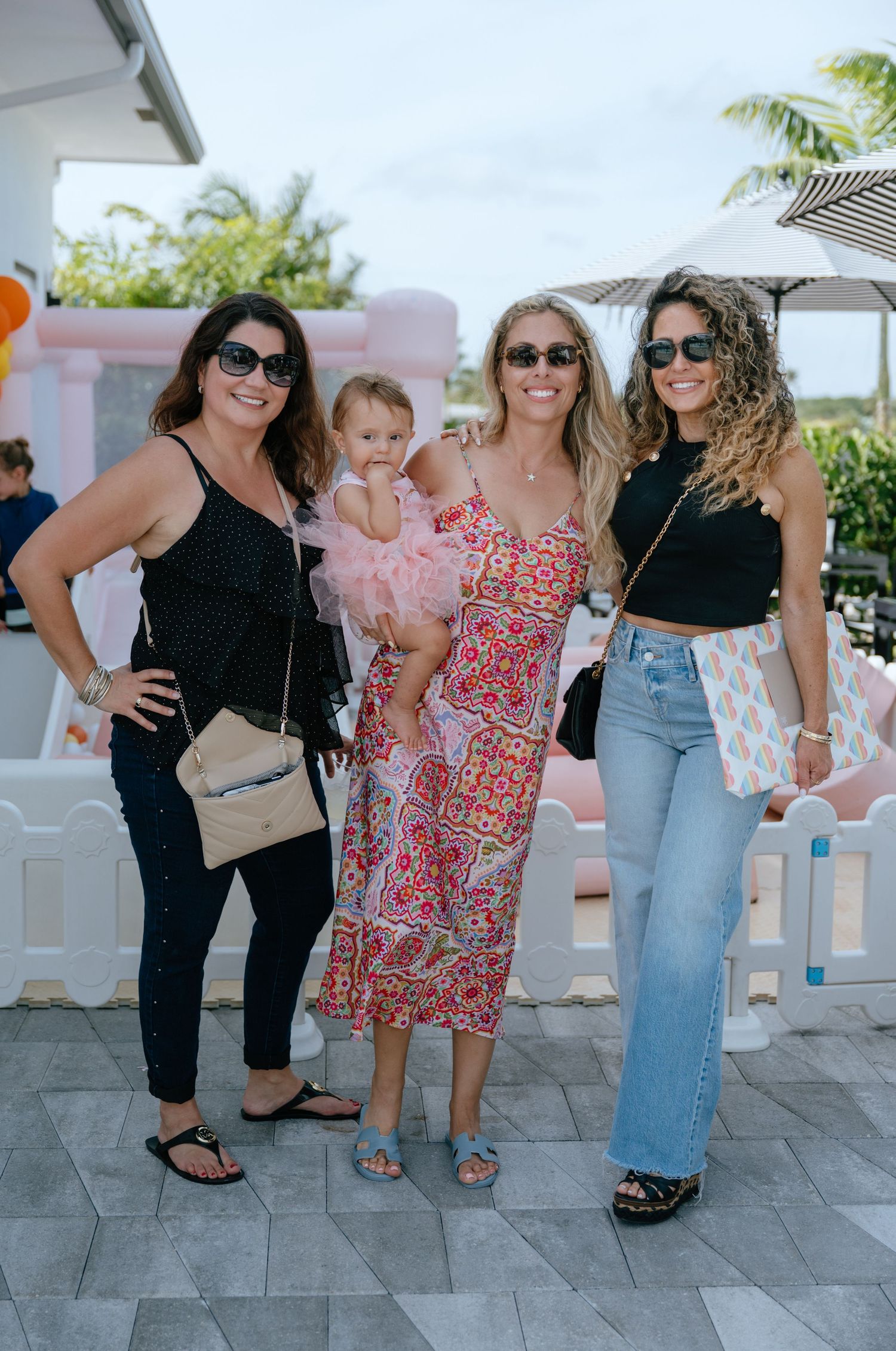 Three women posing together outdoors at a party, one holding a baby dressed in a pink tutu.