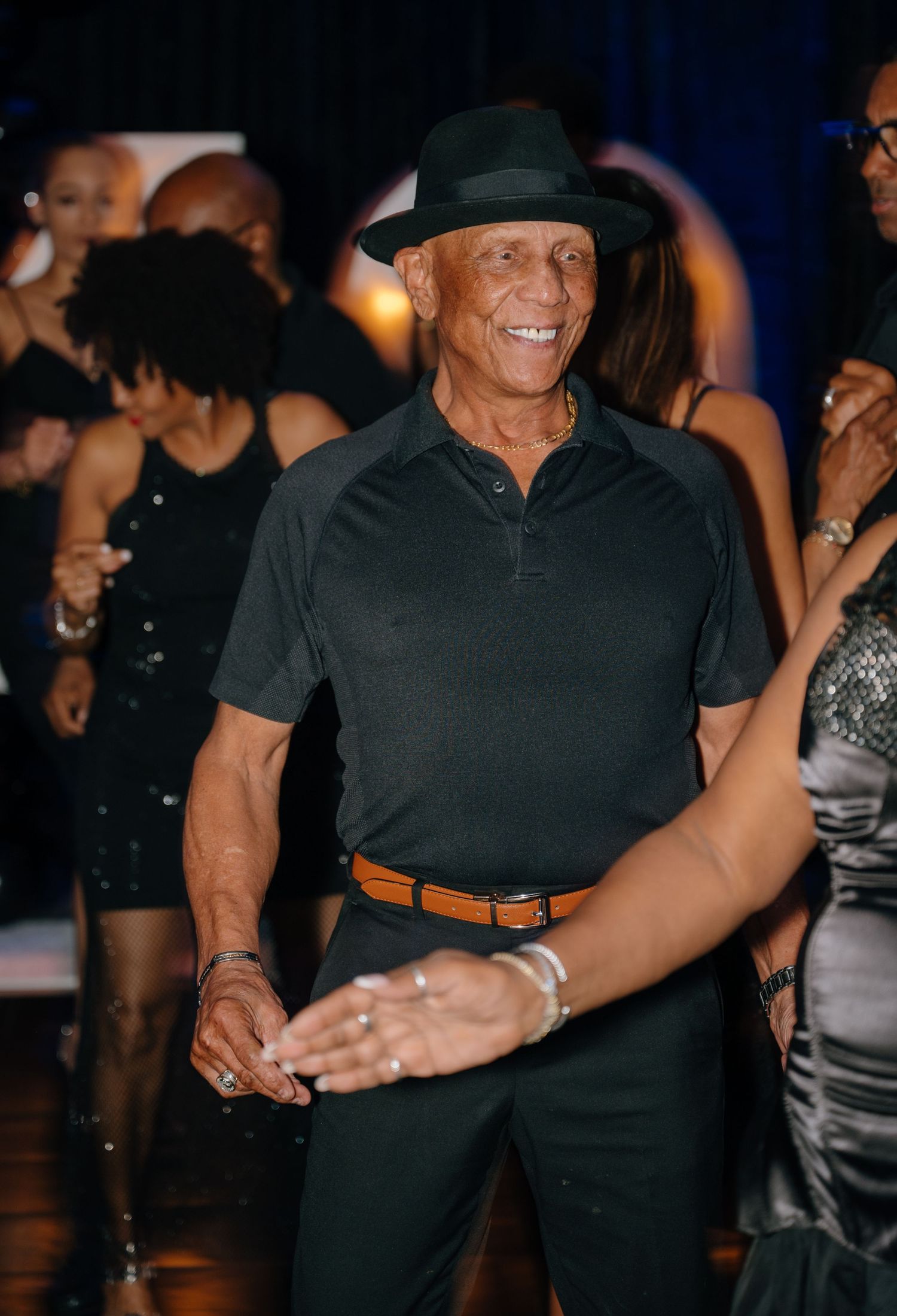 Energetic older man in black outfit and fedora hat dancing at a lively party event.