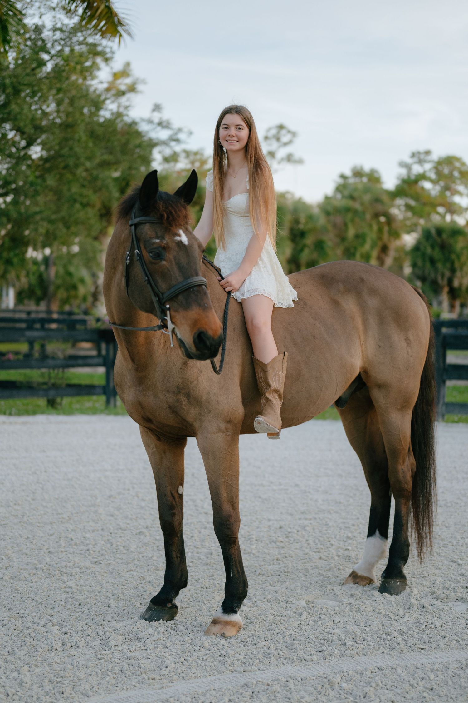 Smiling young woman in white dress and cowboy boots sits atop a bay horse in a sandy arena.