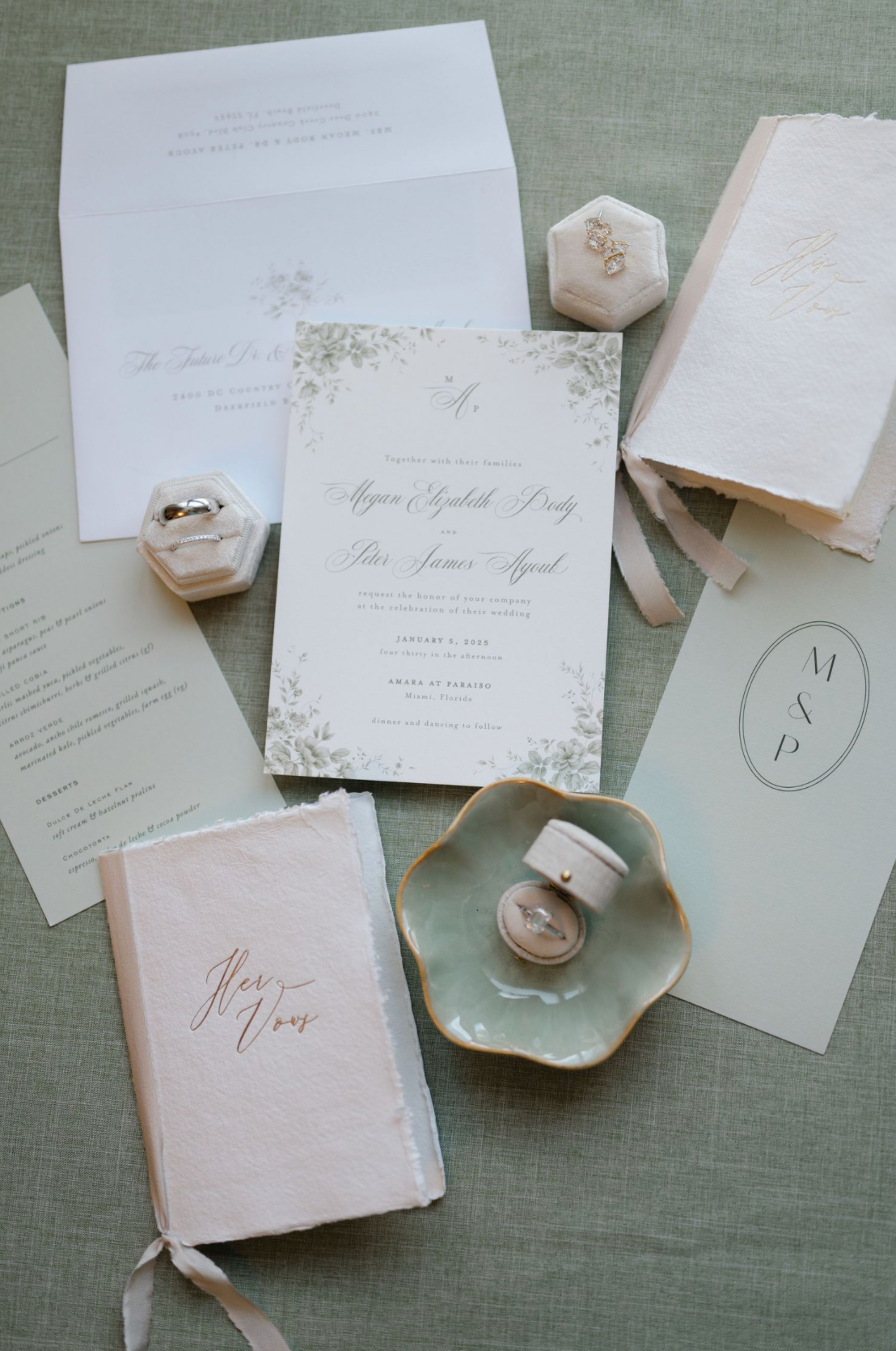 Elegant wedding stationery flatlay featuring floral invitations, velvet ring boxes, vow books, and a ceramic dish.