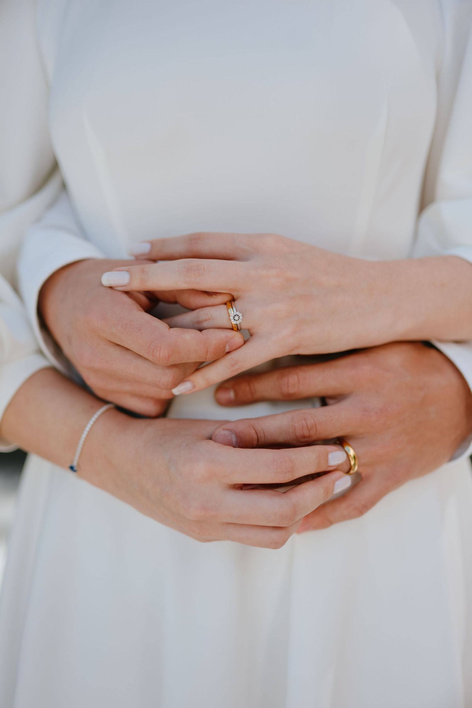 Close-up of newlyweds hands intertwined showing gold wedding rings and diamond engagement ring against a white dress.