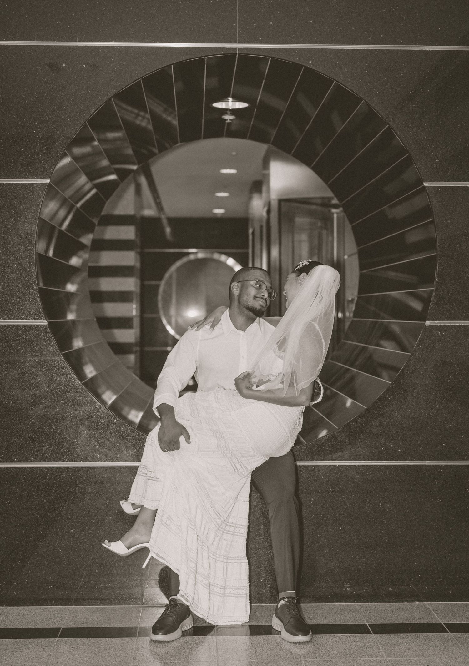 Black and white photo of groom lifting bride in front of a large circular mirror indoors.