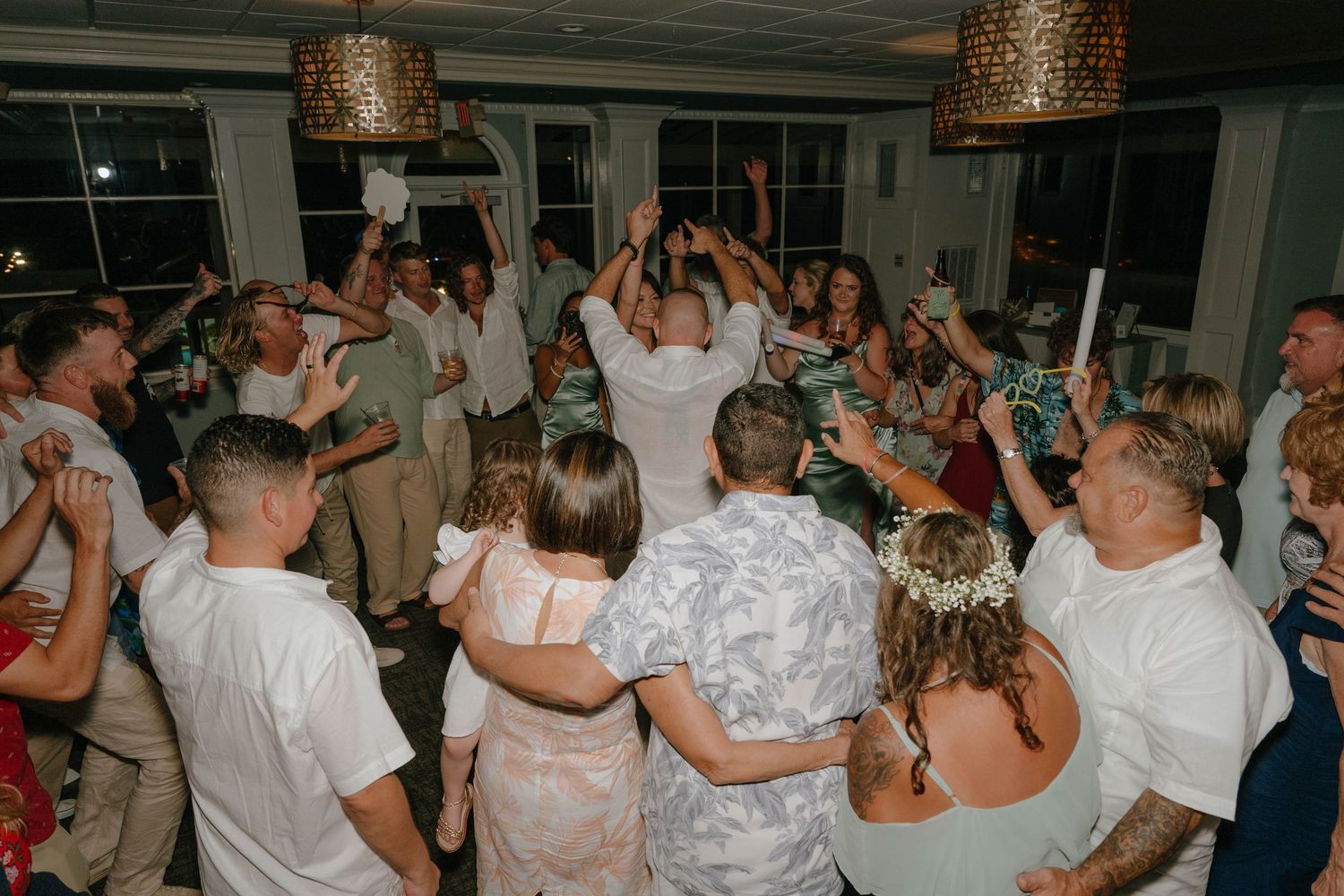 Wedding guests crowd the dance floor celebrating with raised hands and glow sticks at a festive reception.