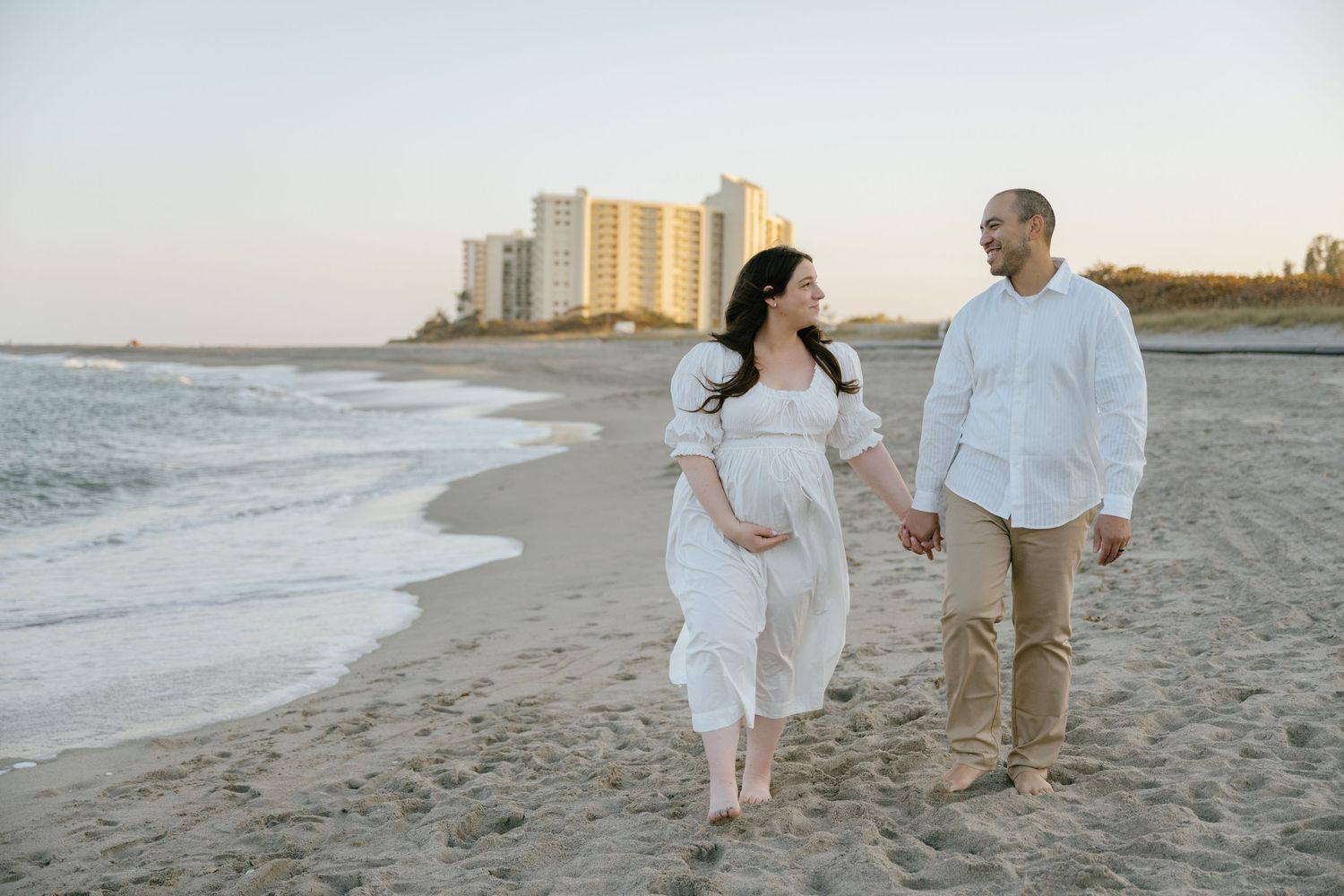 Expecting couple walks hand-in-hand along sandy beach at sunset with city skyline behind.